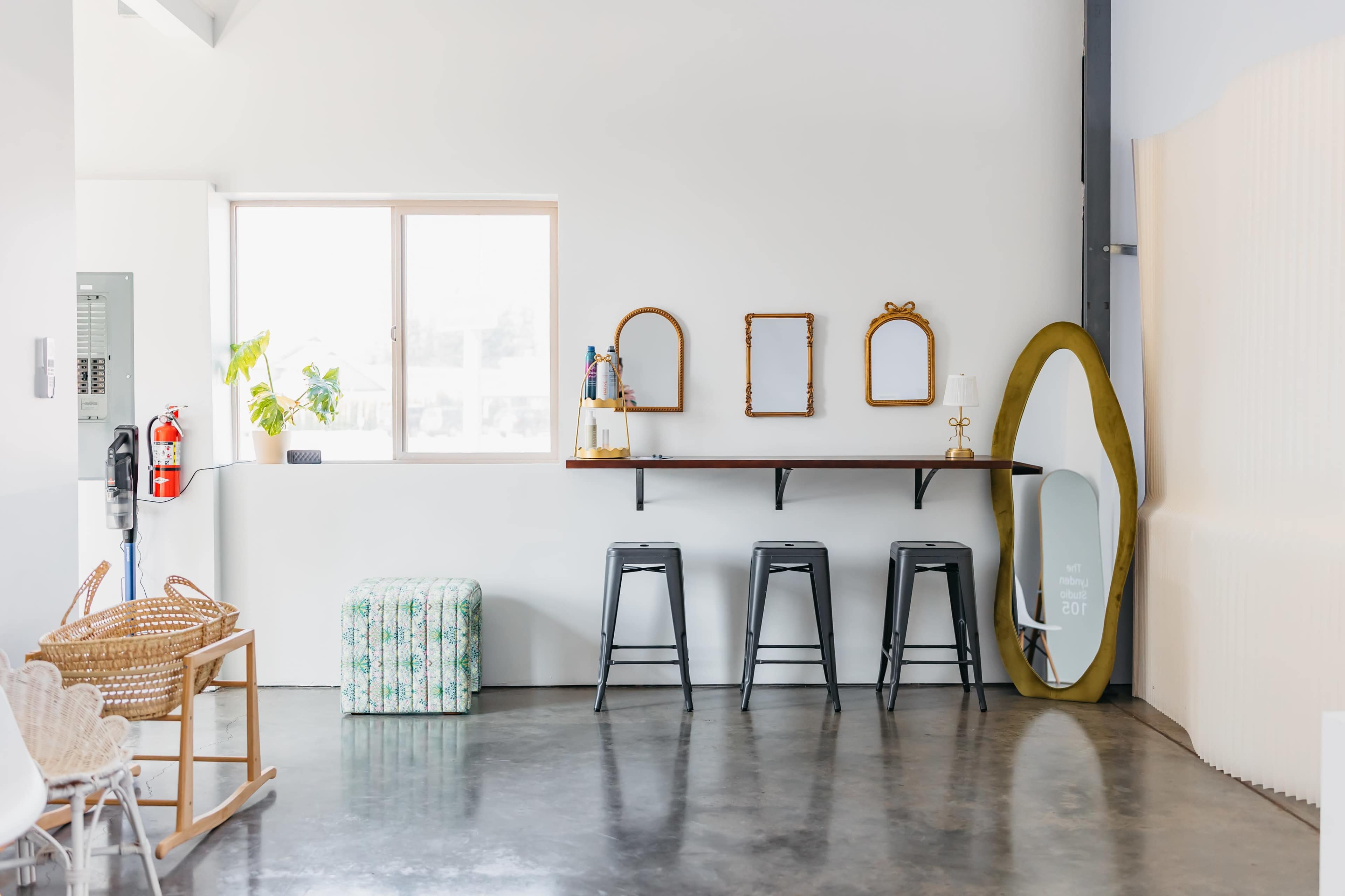 The image shows a minimalist interior with a wooden countertop, three metal stools, a large mirror, and decorative items arranged on a shelf.