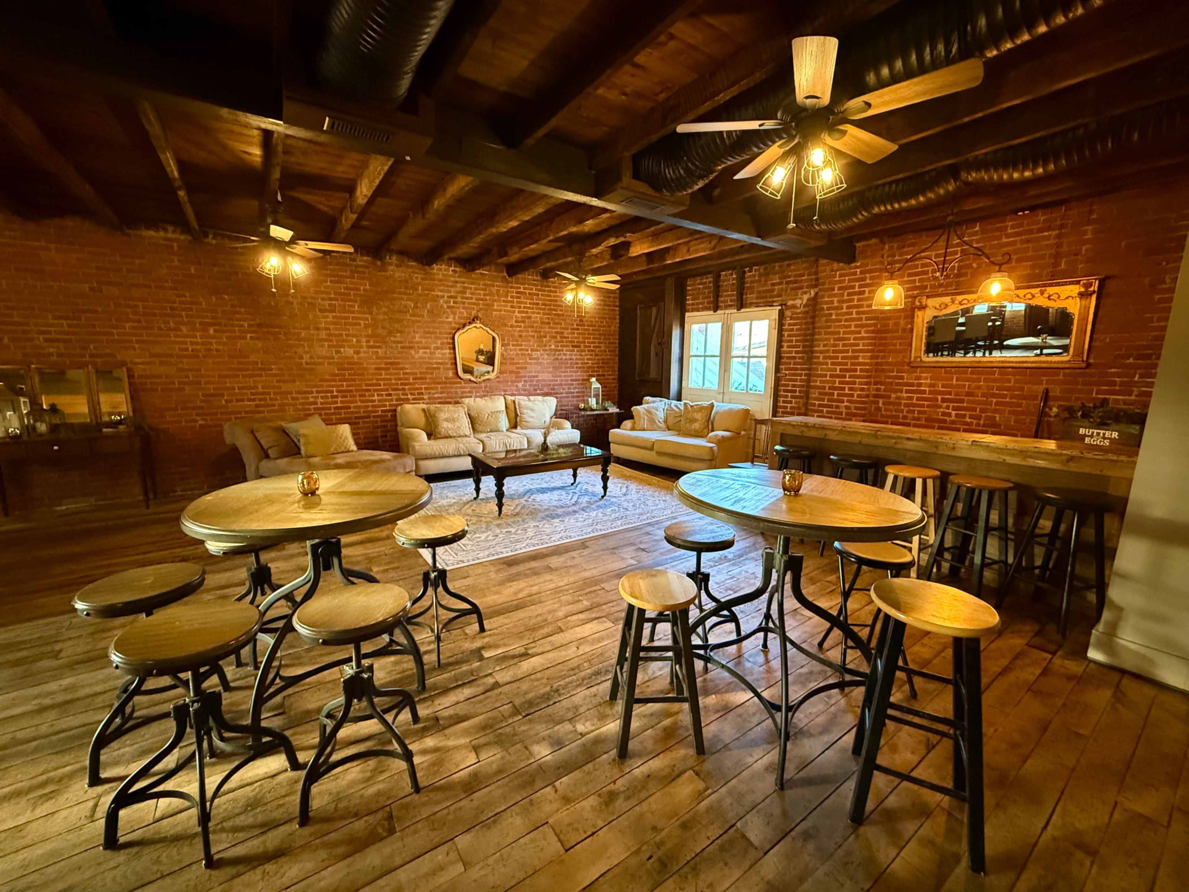 The image shows a rustic interior space with exposed brick walls, wooden flooring, and several seating arrangements, including tables and chairs, well-lit by ceiling fans and warm lighting.