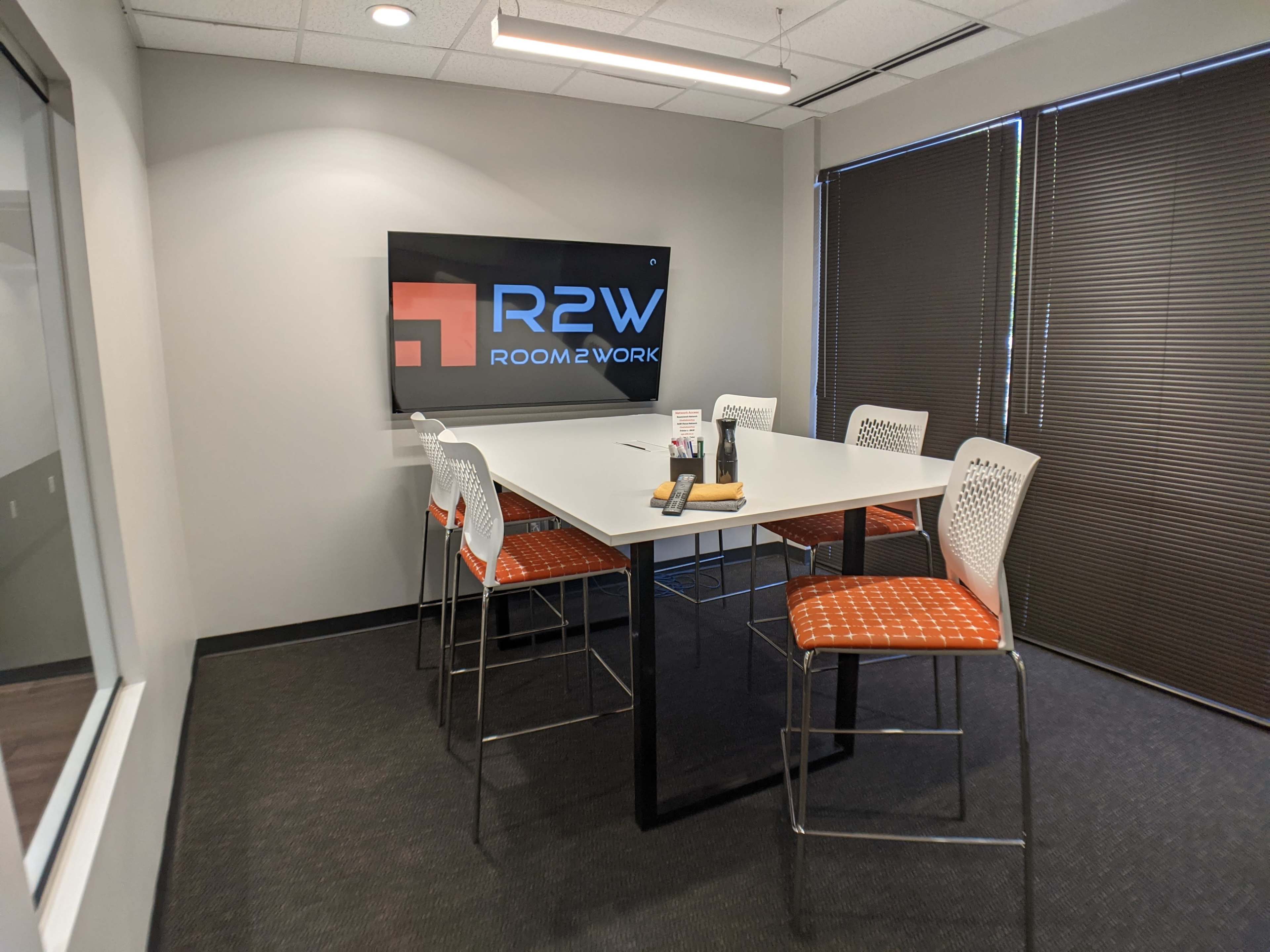A modern meeting room with a rectangular table surrounded by six high-backed stools and a screen displaying the "R2W ROOM2WORK" logo on the wall.