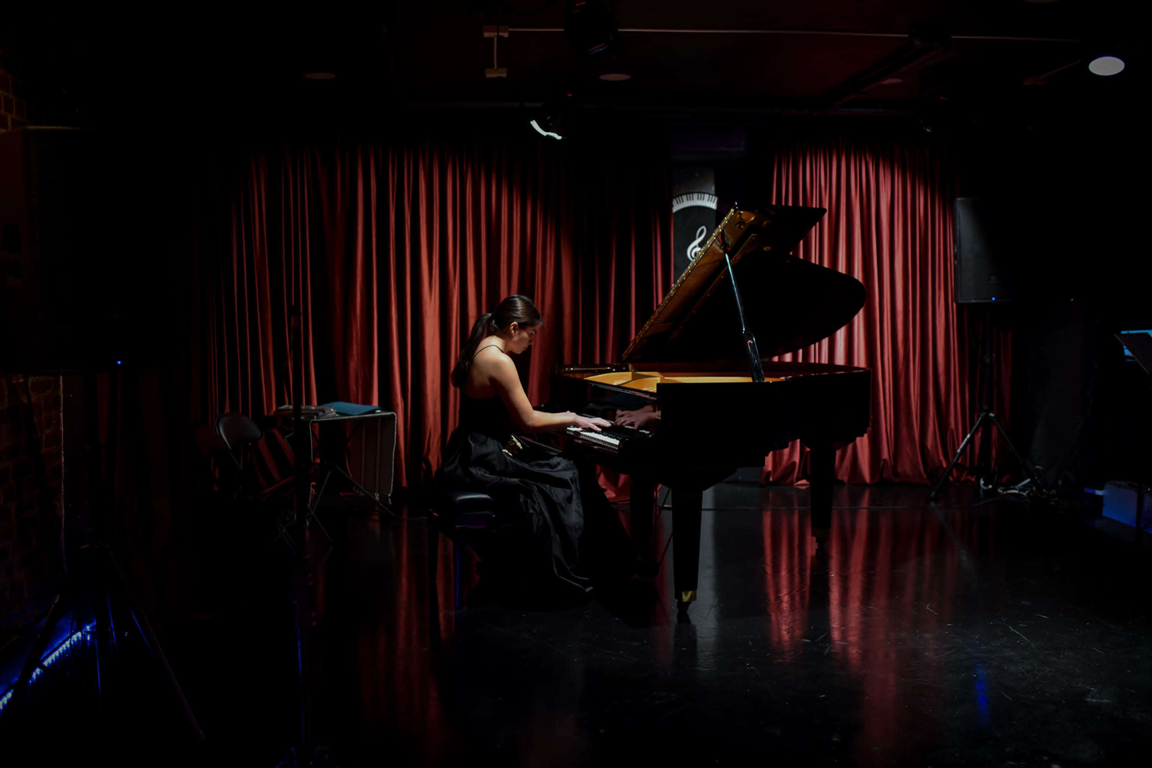 A woman in a black dress plays a grand piano on a dimly lit stage with red curtains.