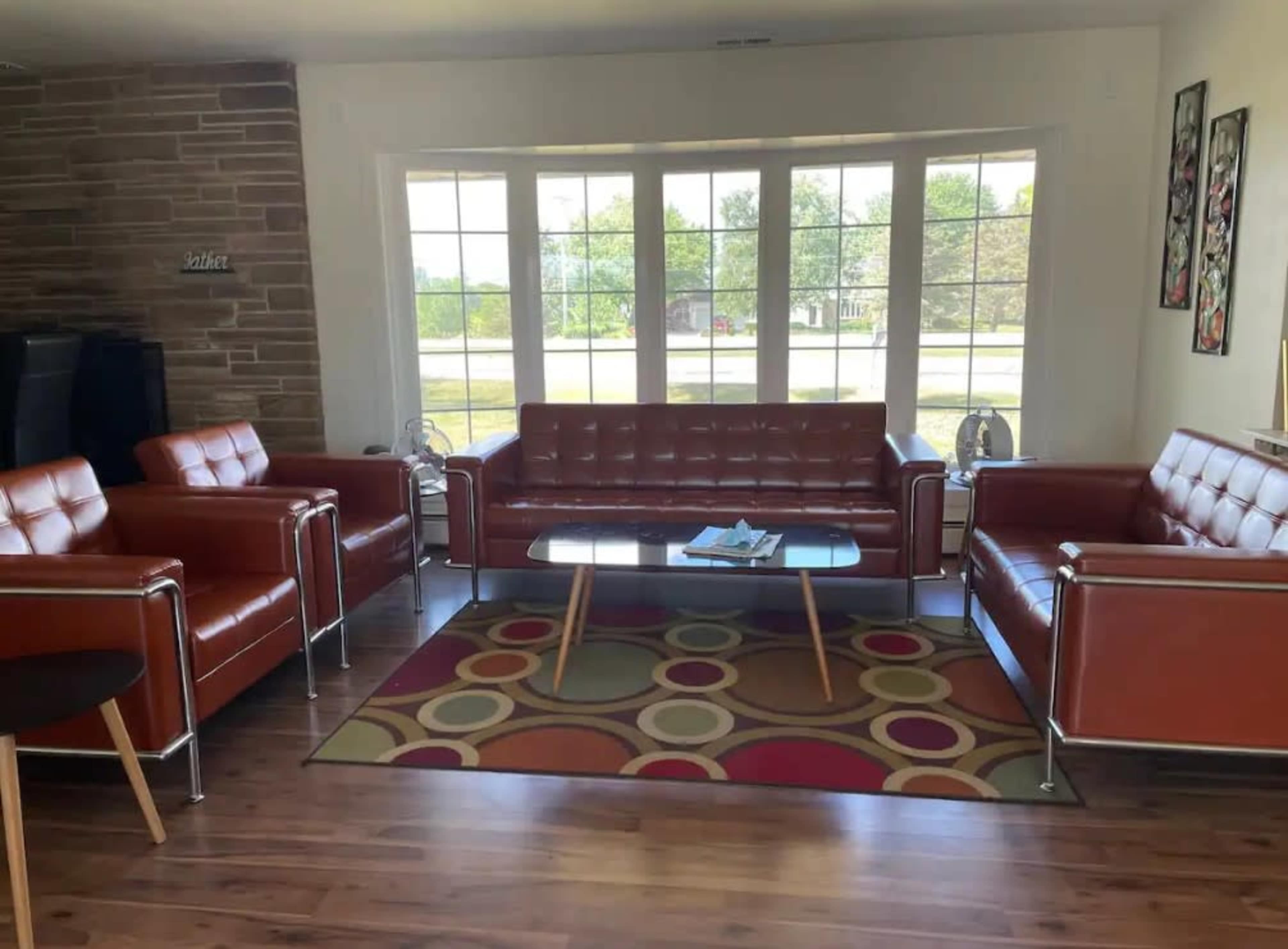 A modern living room featuring brown leather sofas and armchairs, a glass coffee table, and a colorful circular patterned rug, all positioned near large windows.