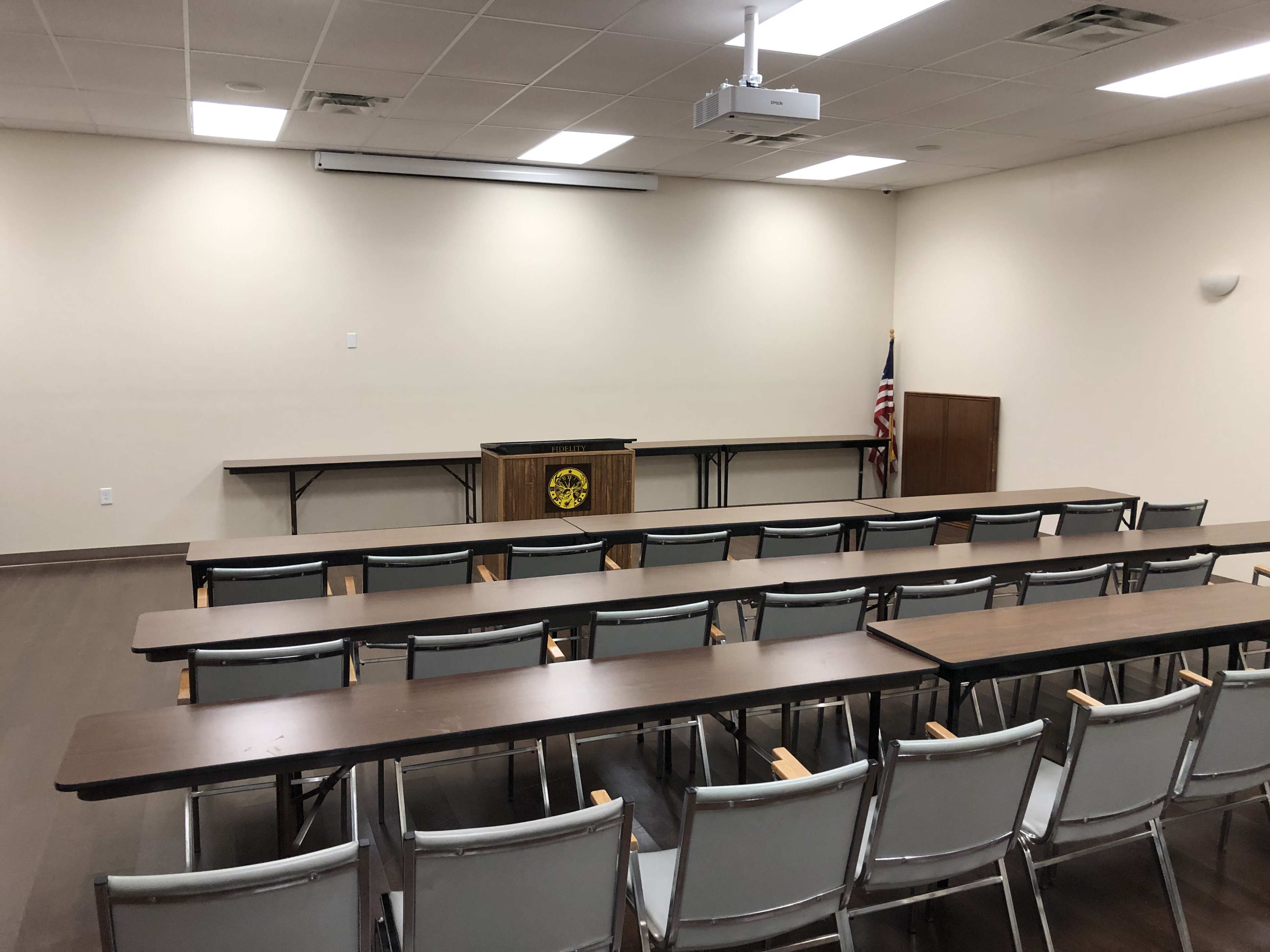 A conference room with several tables arranged in rows and chairs facing a podium, featuring an American flag in the corner.