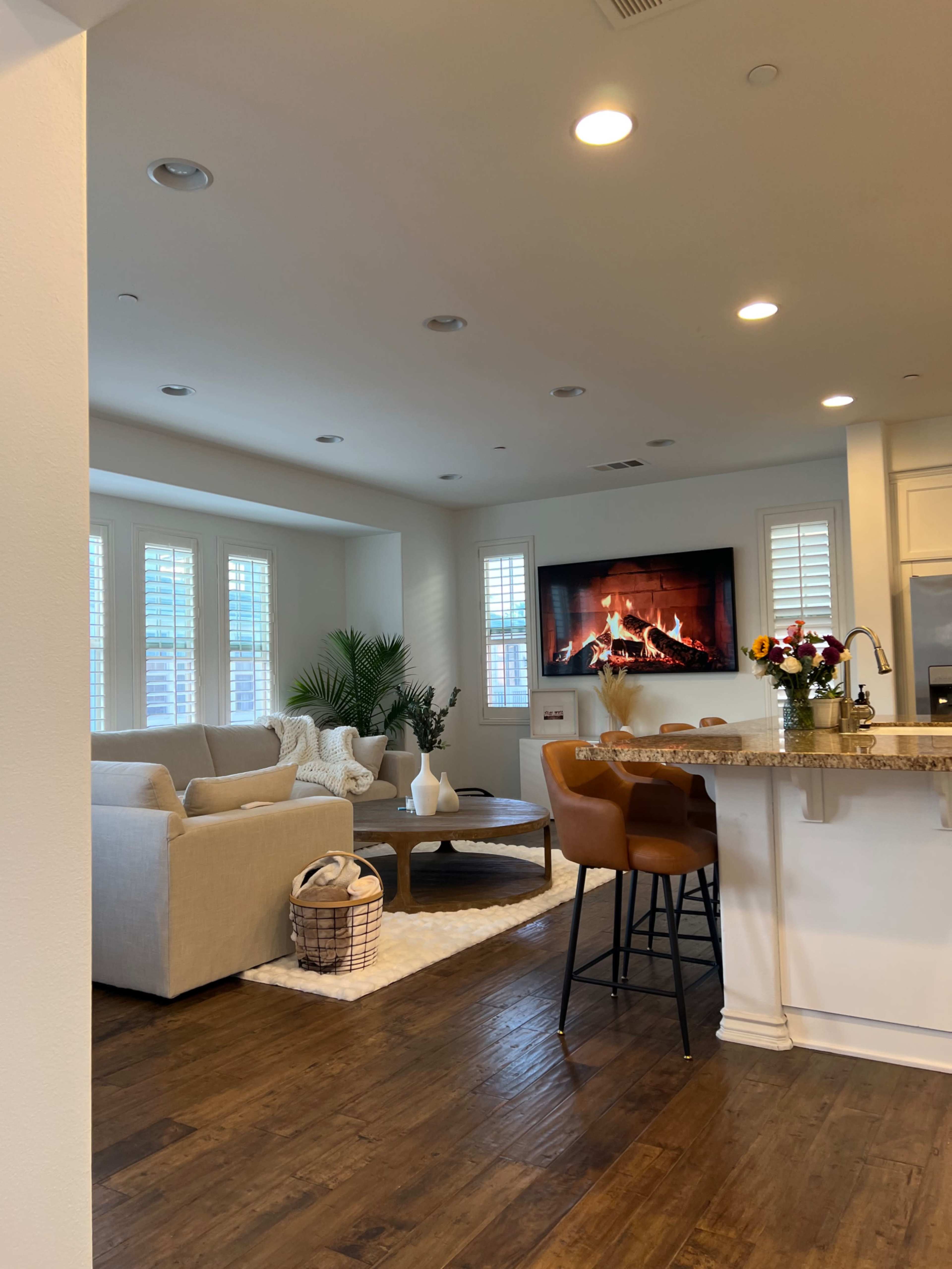 A modern living room features a light-colored sofa, a round coffee table, and a wall-mounted television displaying a fireplace, with a kitchen bar in the foreground.