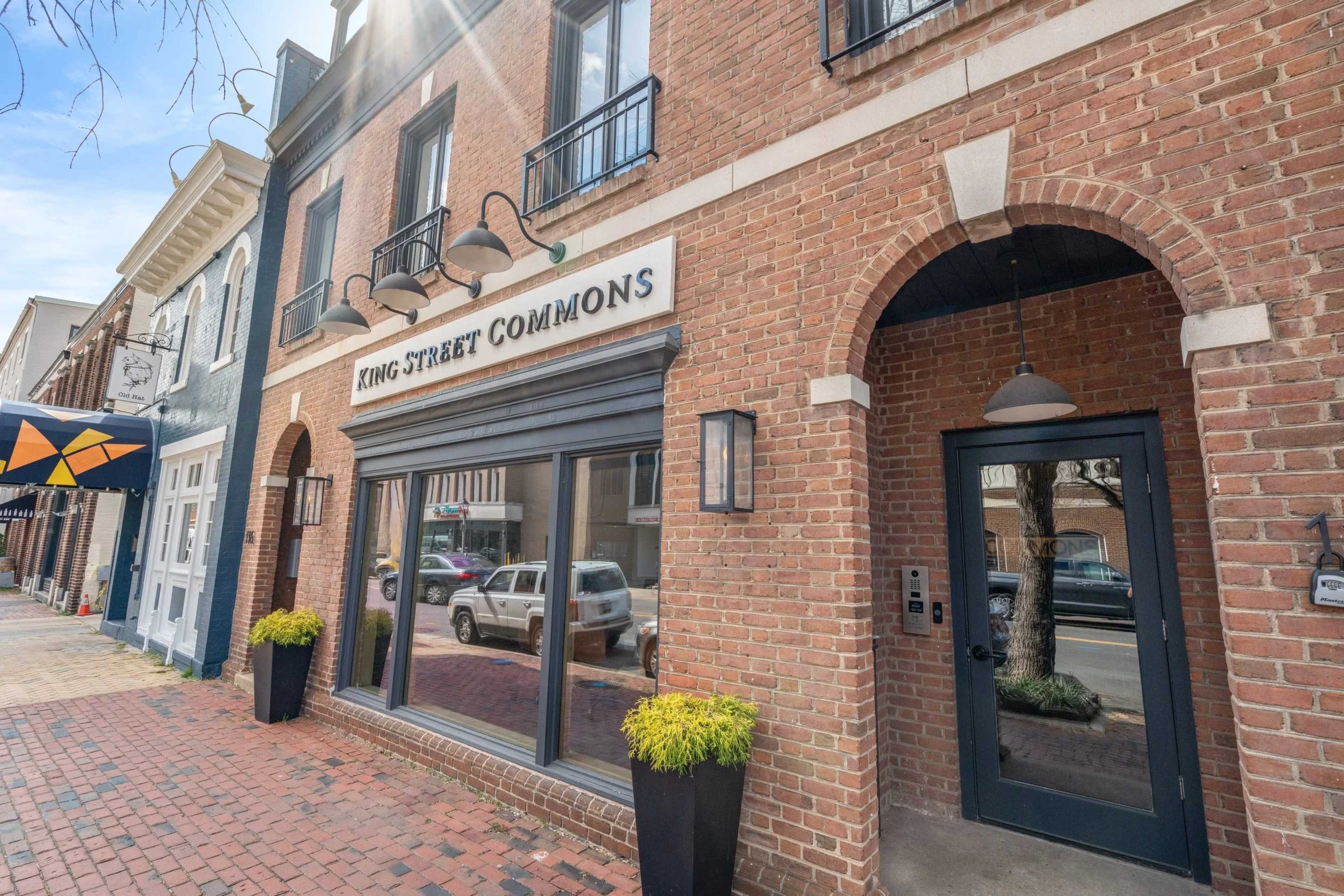 The image shows the brick facade of a building with a sign reading "KING STREET COMMONS" above large windows and a front door, situated on a street with trees and parked cars.