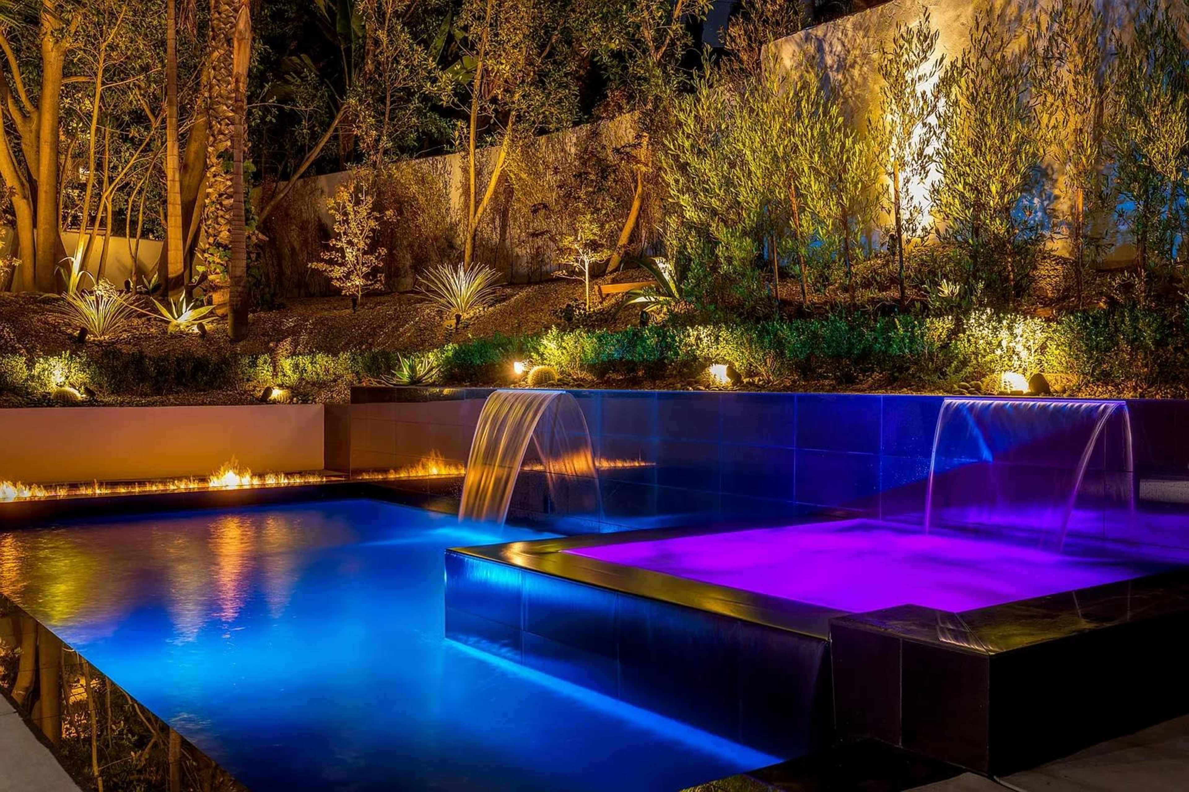 The image shows a modern outdoor pool area at night, featuring illuminated water features and colorful lighting.