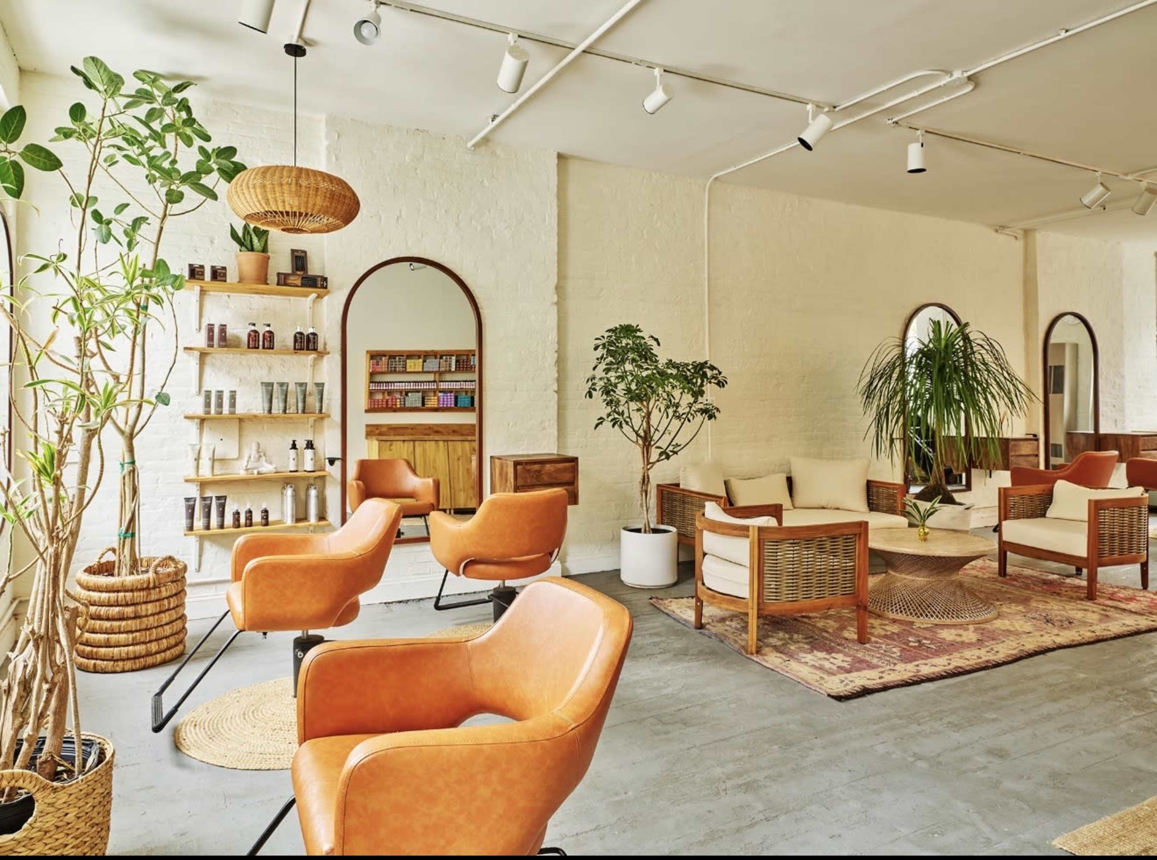 The image shows a modern, airy interior of a wellness or beauty studio with orange armchairs, wooden shelving, a variety of plants, and large mirrors on the walls.