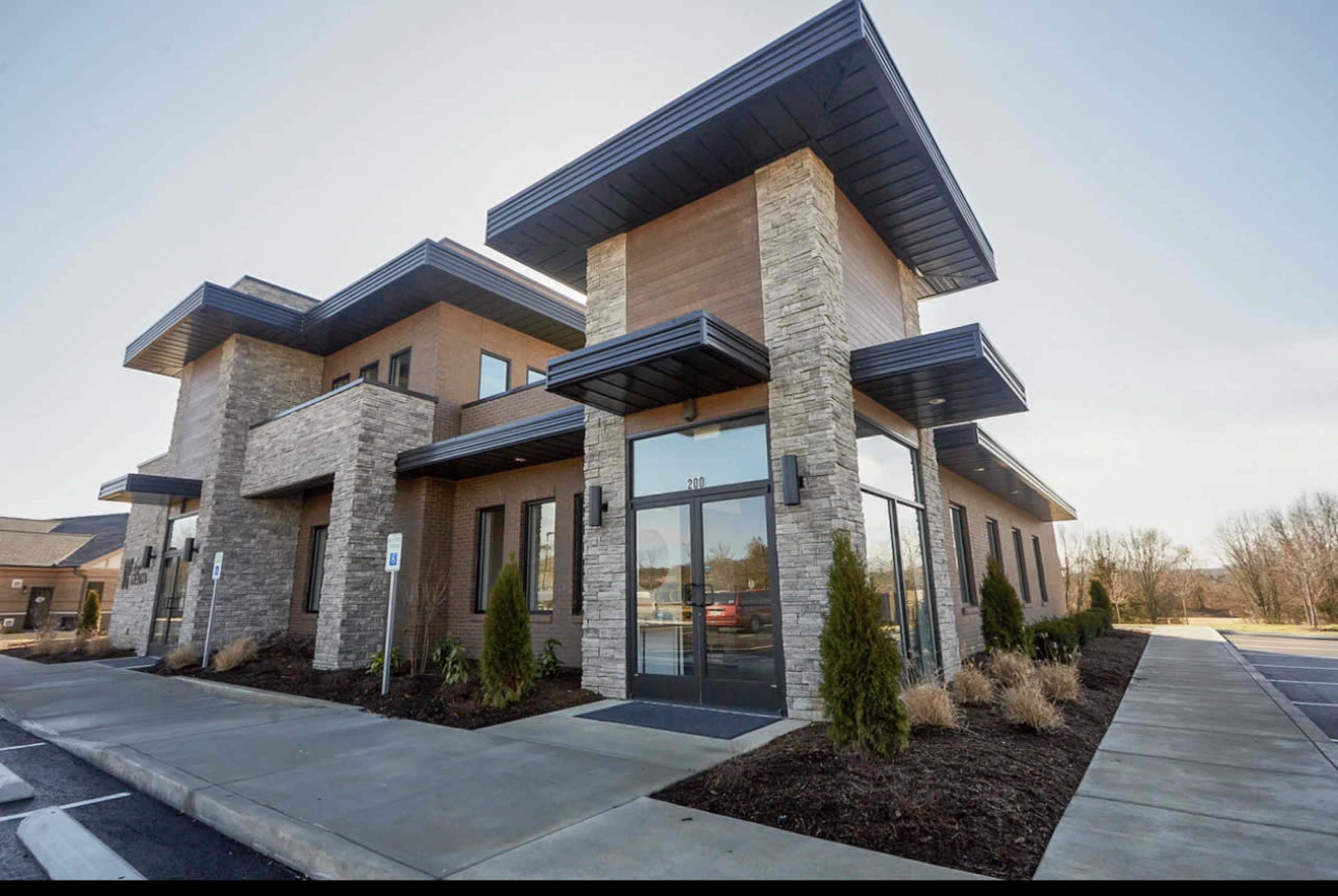 A modern commercial building with a stone and wood facade, featuring large glass doors and landscaping in front.