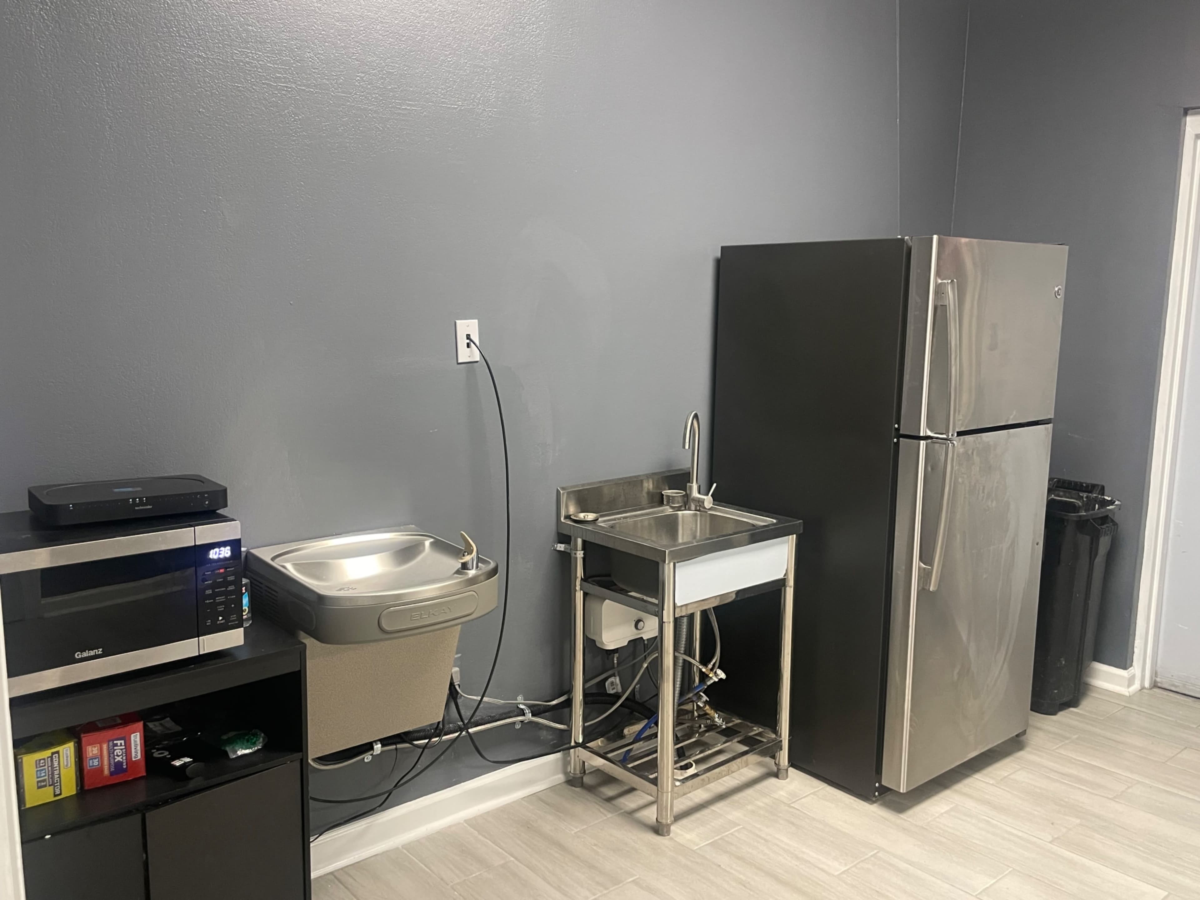 A kitchen area with a stainless steel sink, a refrigerator, a microwave, and a waste bin.