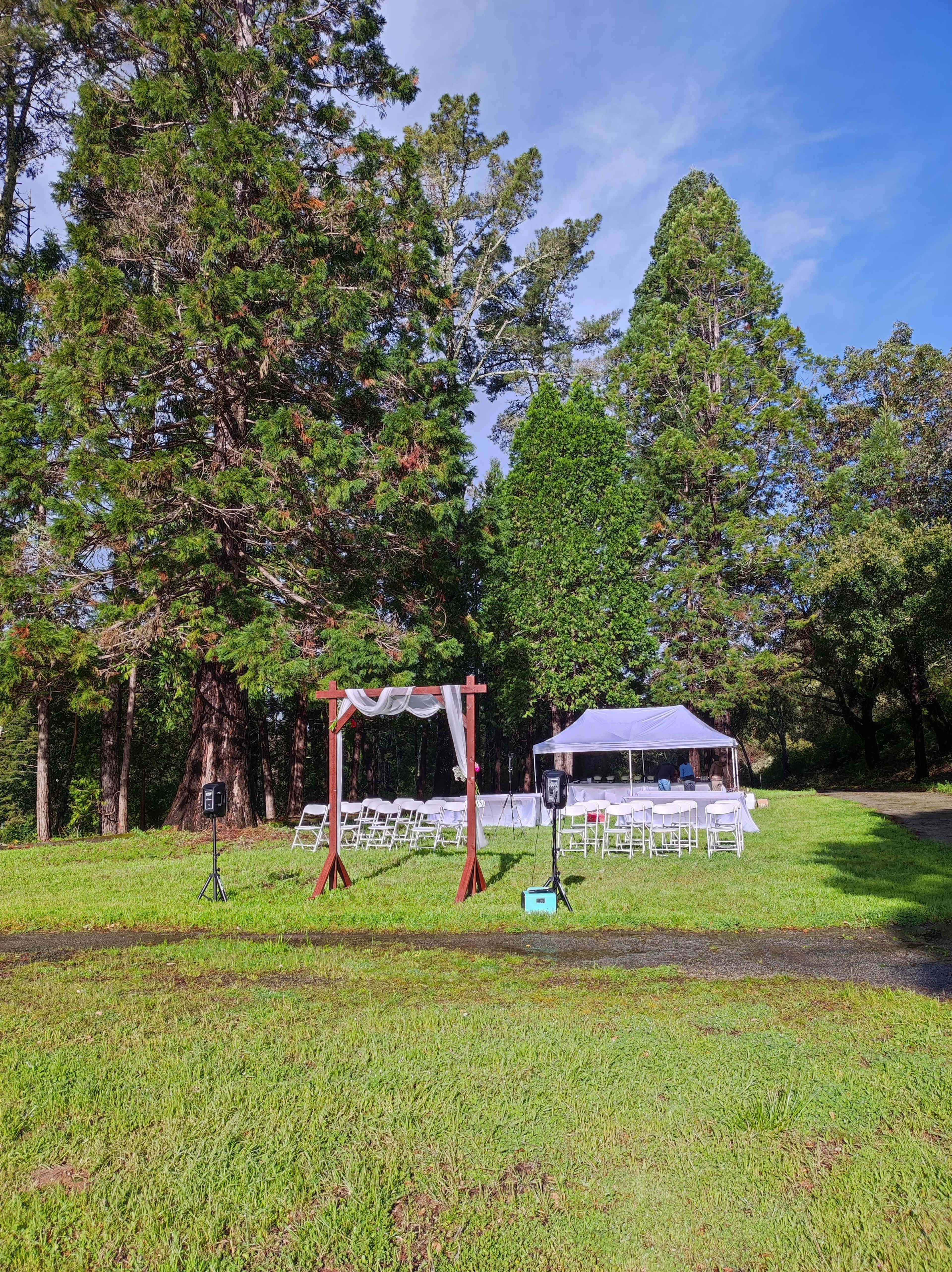 A wedding setup is arranged outdoors with a wooden arch, white chairs, and a tent surrounded by tall trees.