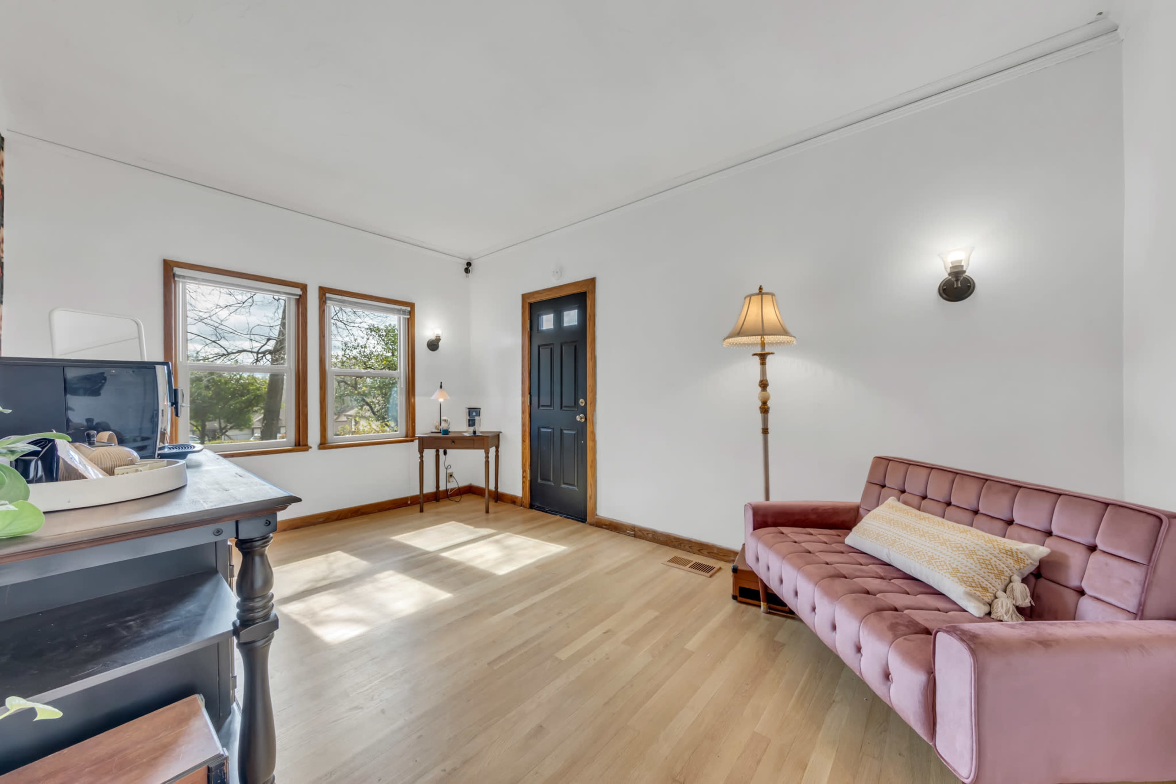 The room features a pink sofa, a wooden table with a lamp, and large windows allowing natural light to illuminate the space.