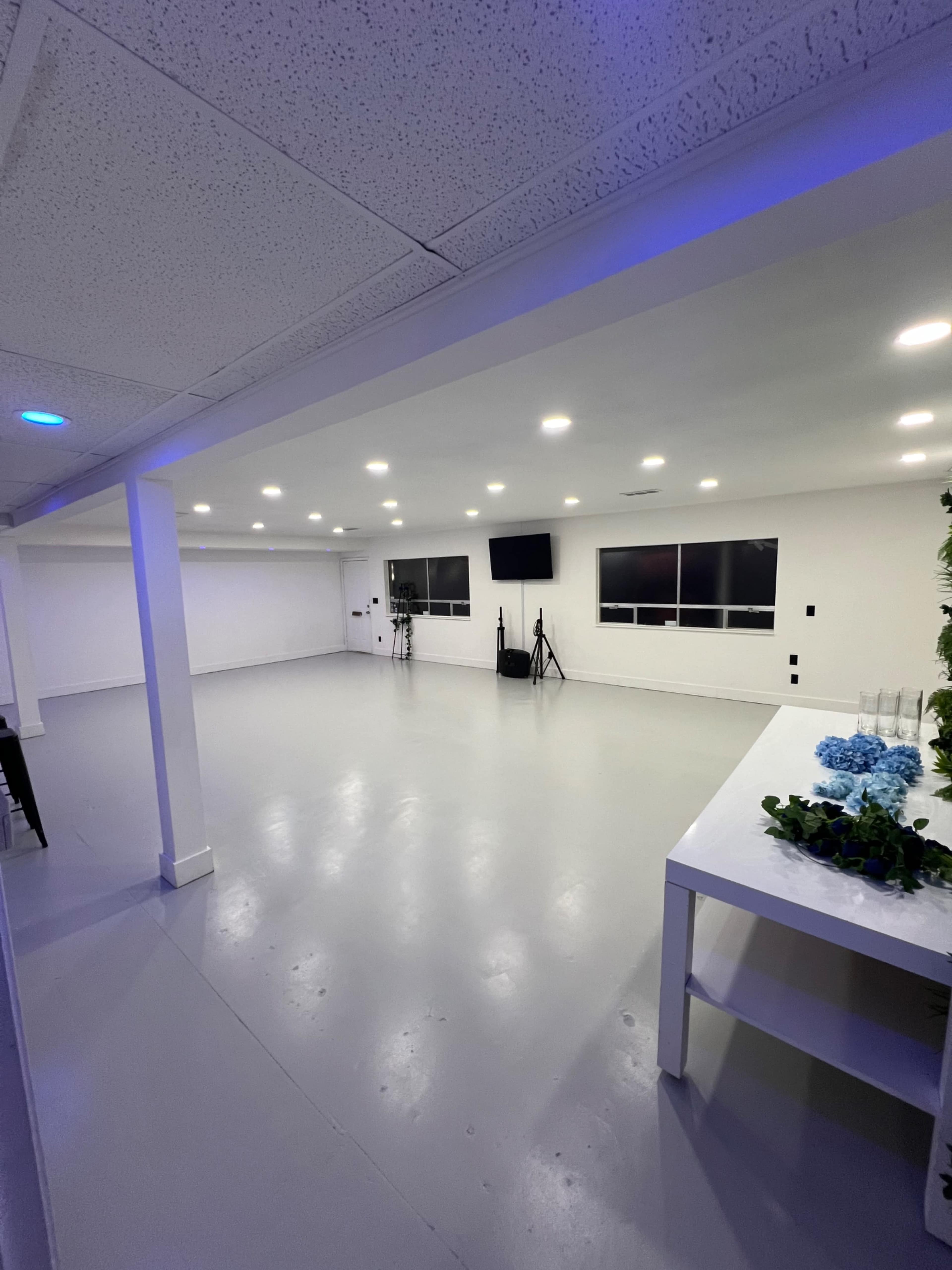 The image shows a spacious, well-lit room with gray flooring, white walls, and two televisions mounted on the walls.