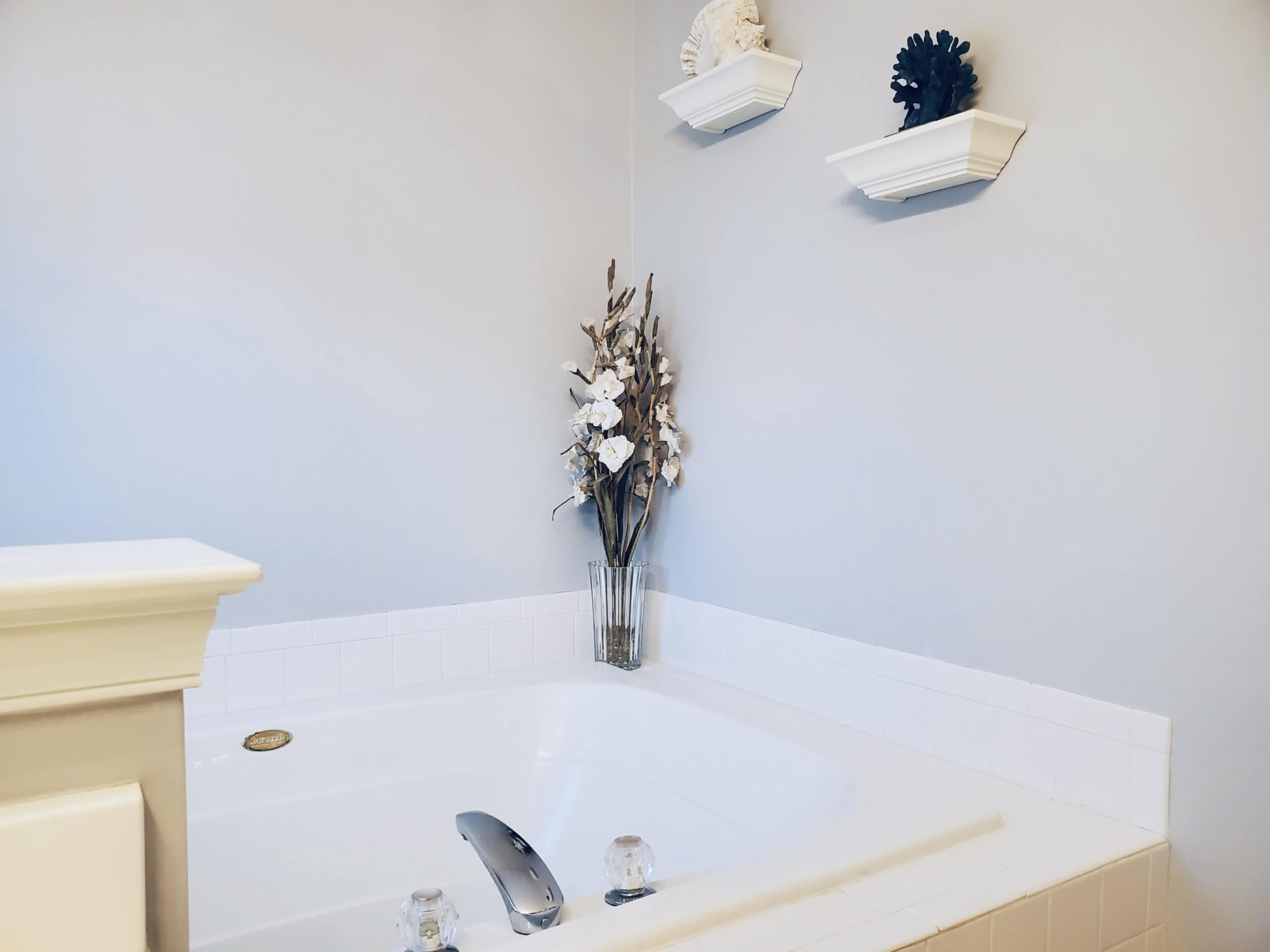 A corner of a bathroom featuring a white jetted tub accompanied by a clear vase with decorative flowers and two wall-mounted shelves.