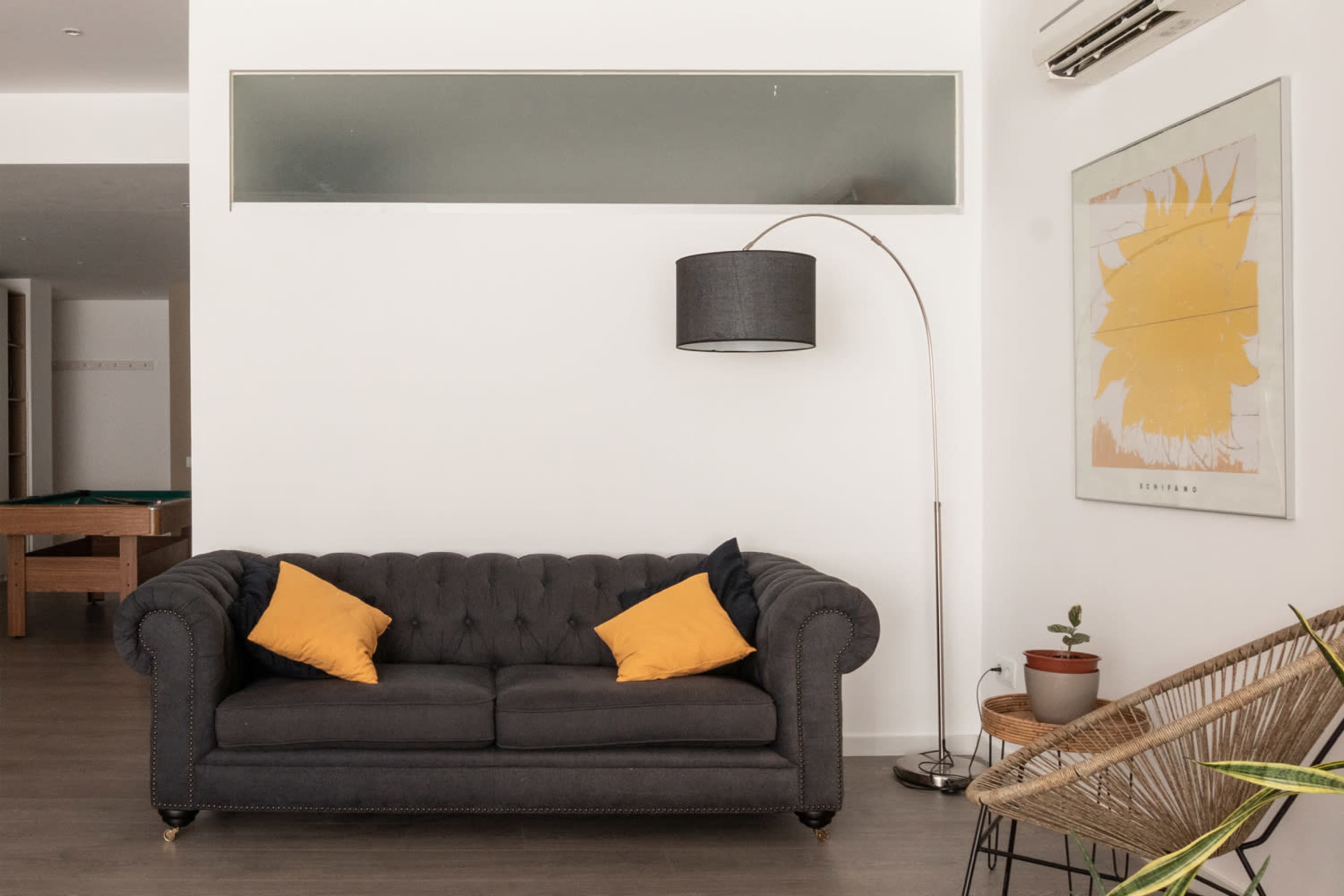 The image shows a modern living room featuring a gray tufted sofa with yellow pillows, a floor lamp, a decorative plant, and a pool table in the background.