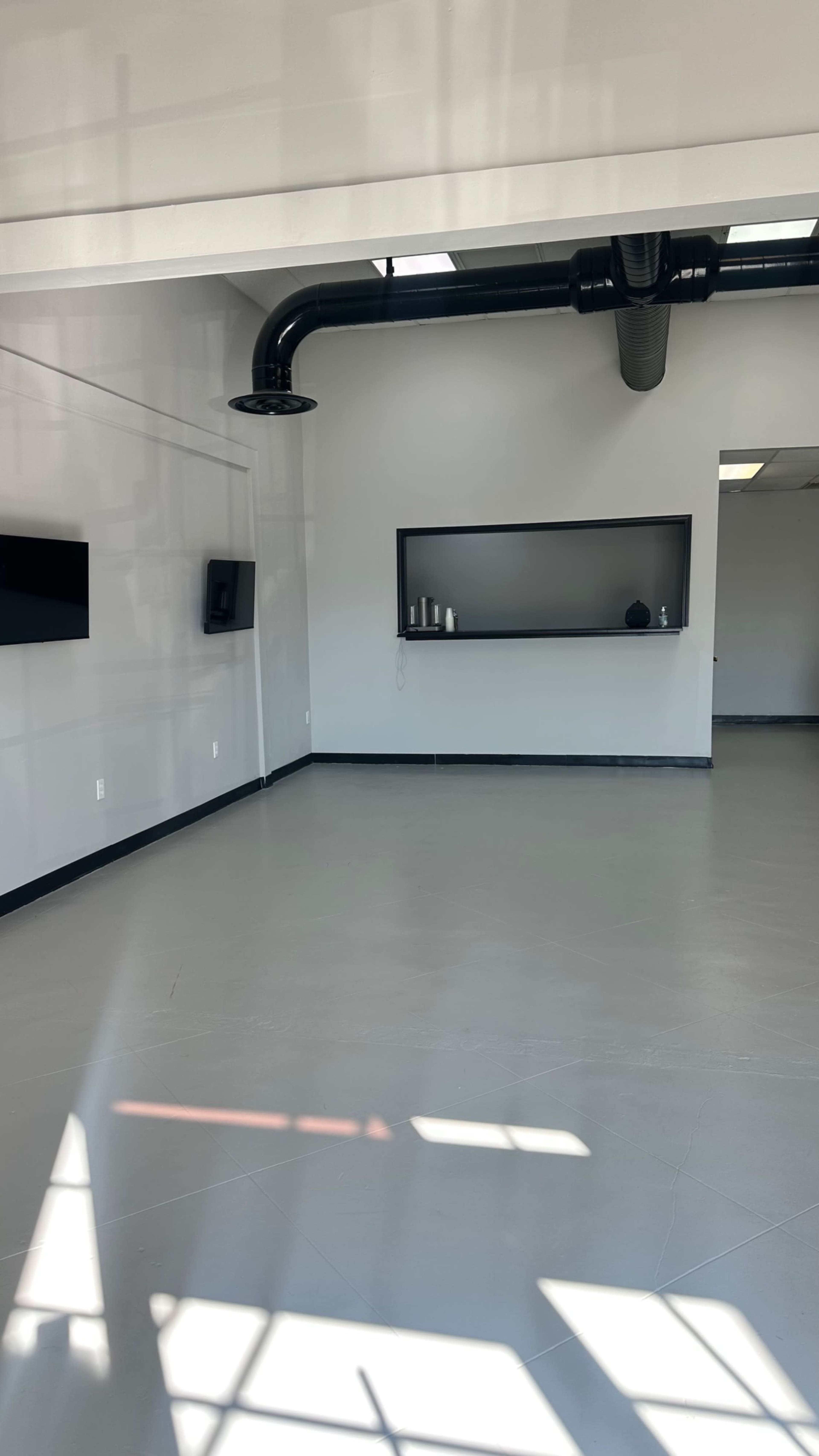 The image shows a spacious, empty room with gray flooring, high ceilings, and two mounted screens on the walls, along with a shelf in the background.