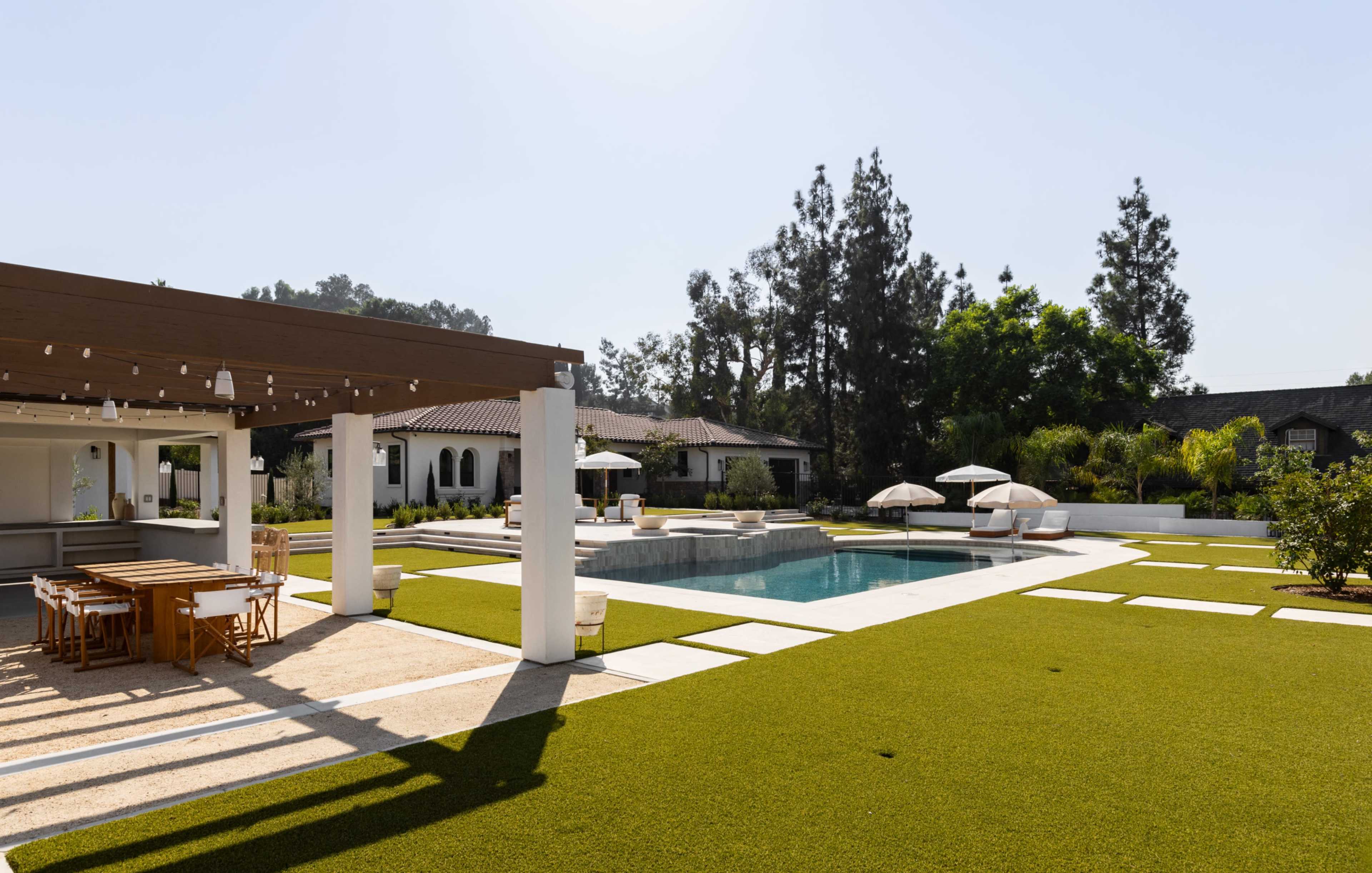 The image shows a modern outdoor space featuring a swimming pool, sun loungers, and a dining area under a pergola surrounded by landscaped greenery.