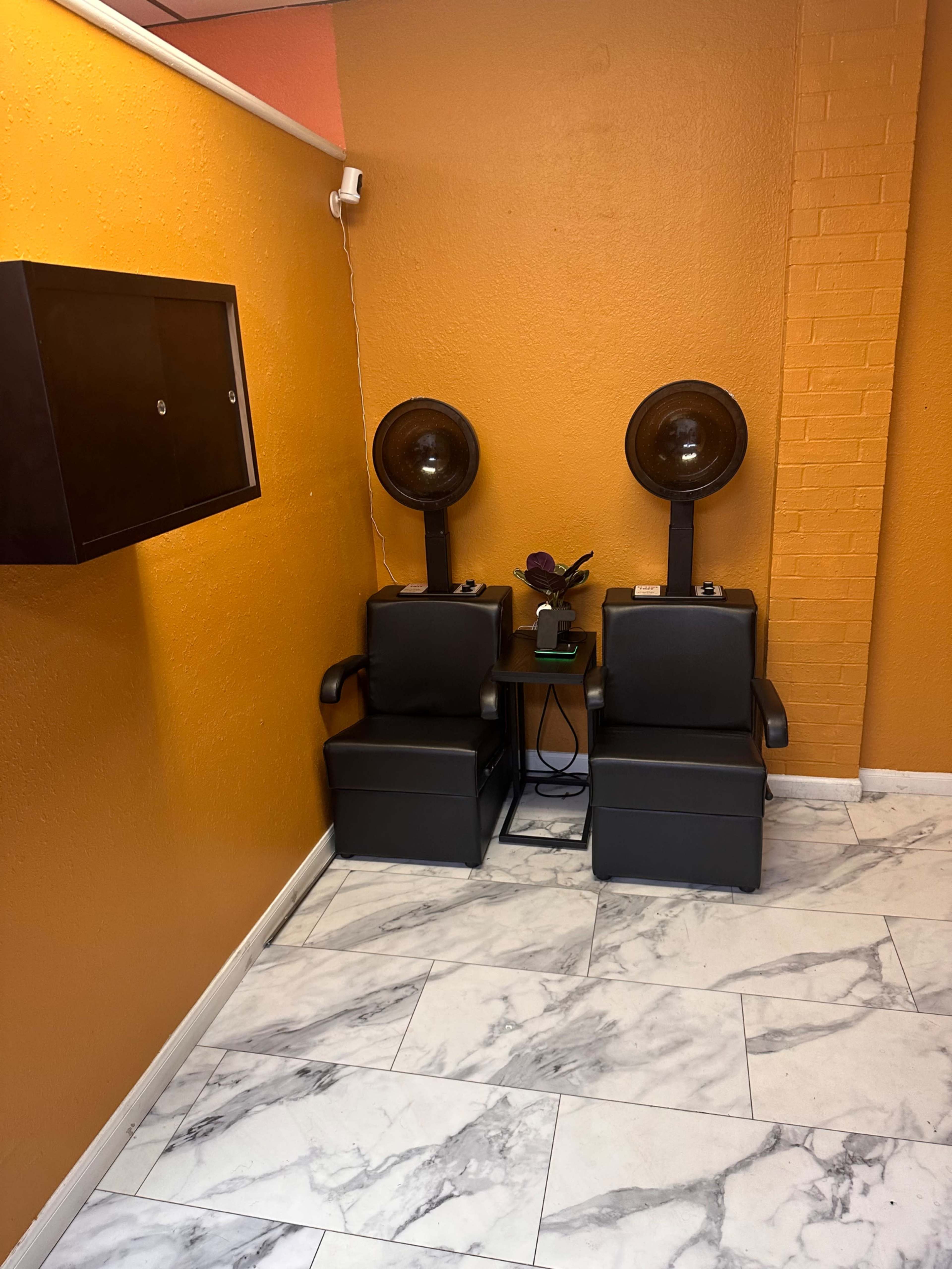 Two black salon chairs with hooded hair dryers are positioned against an orange wall in a room with marble tile flooring.