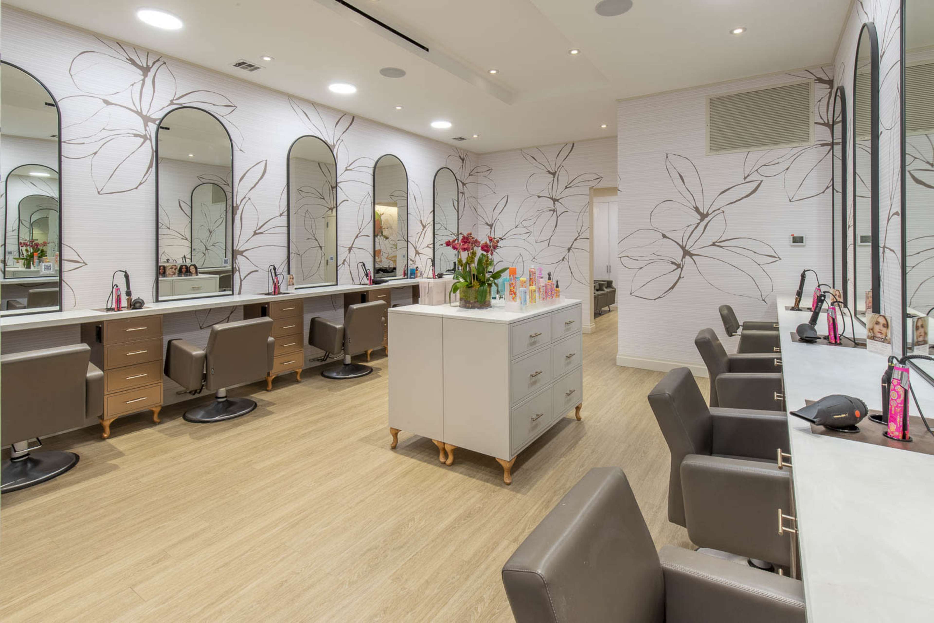 The image shows a modern beauty salon interior with several styling stations featuring large mirrors, light-colored walls adorned with floral patterns, and a central counter.