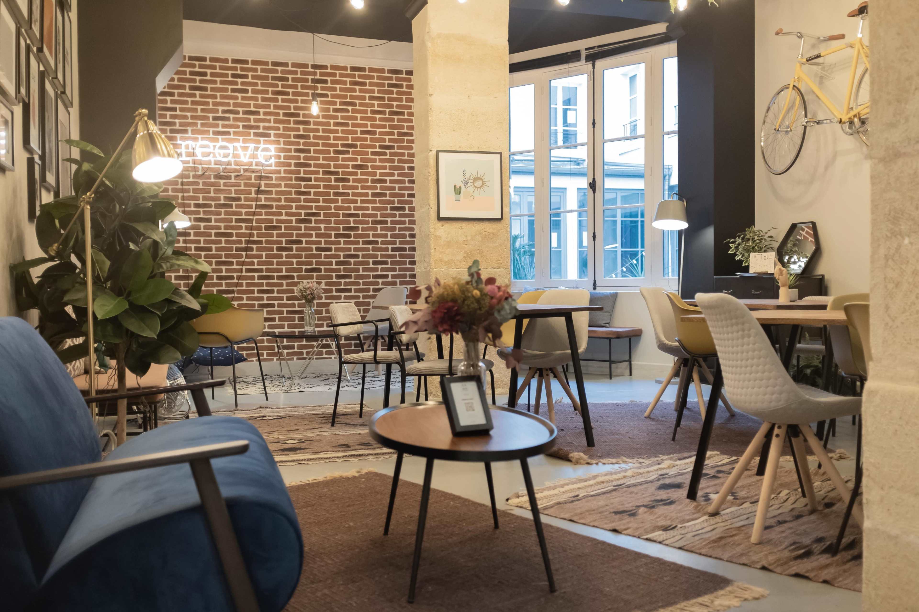 The image depicts a cozy café with a mix of seating arrangements, including blue sofas and wooden tables, set against a backdrop of exposed brick walls and large windows.