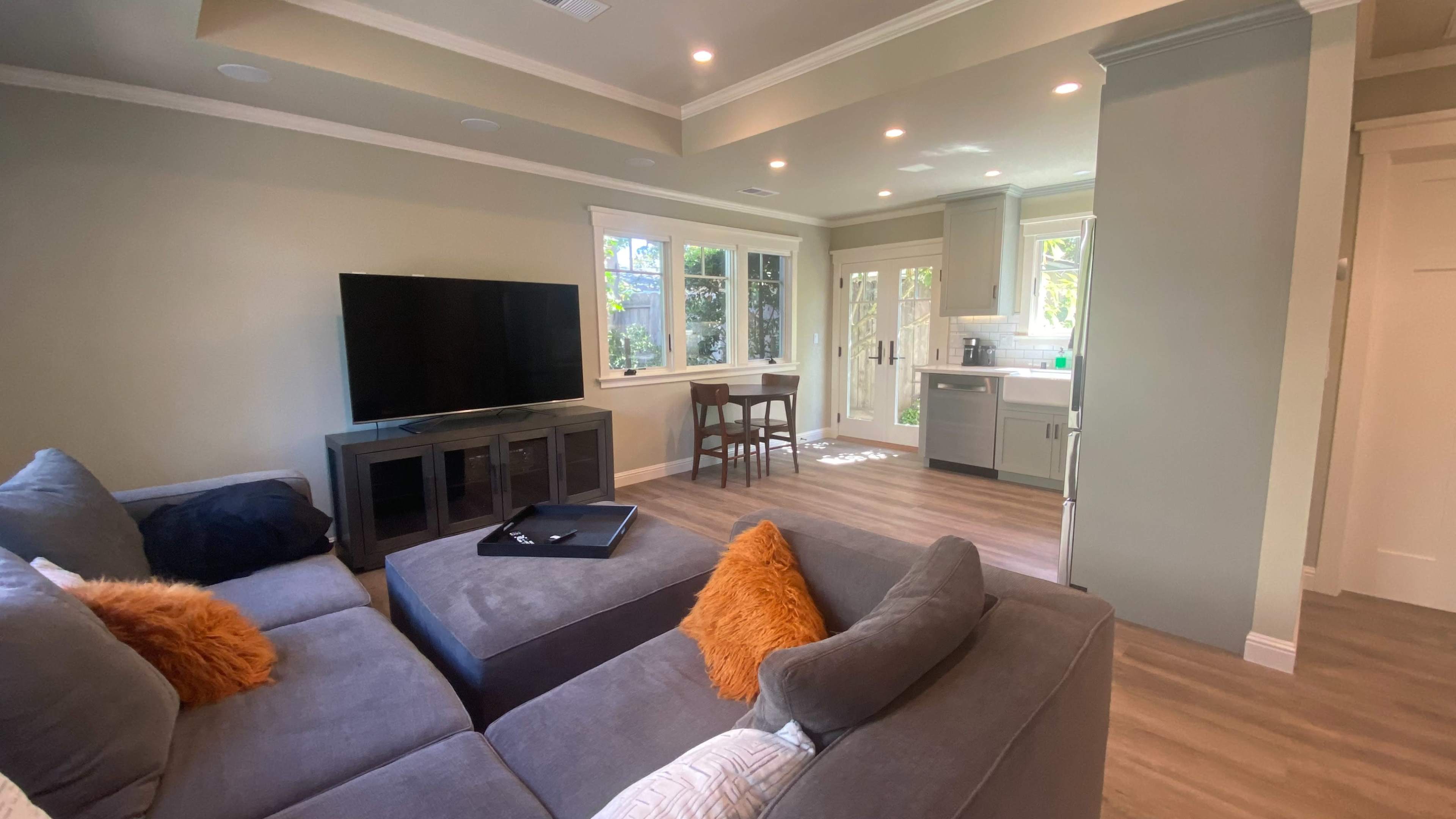 The image shows a modern living room area with a sectional sofa, a television unit, and a small dining space adjacent to a kitchen.