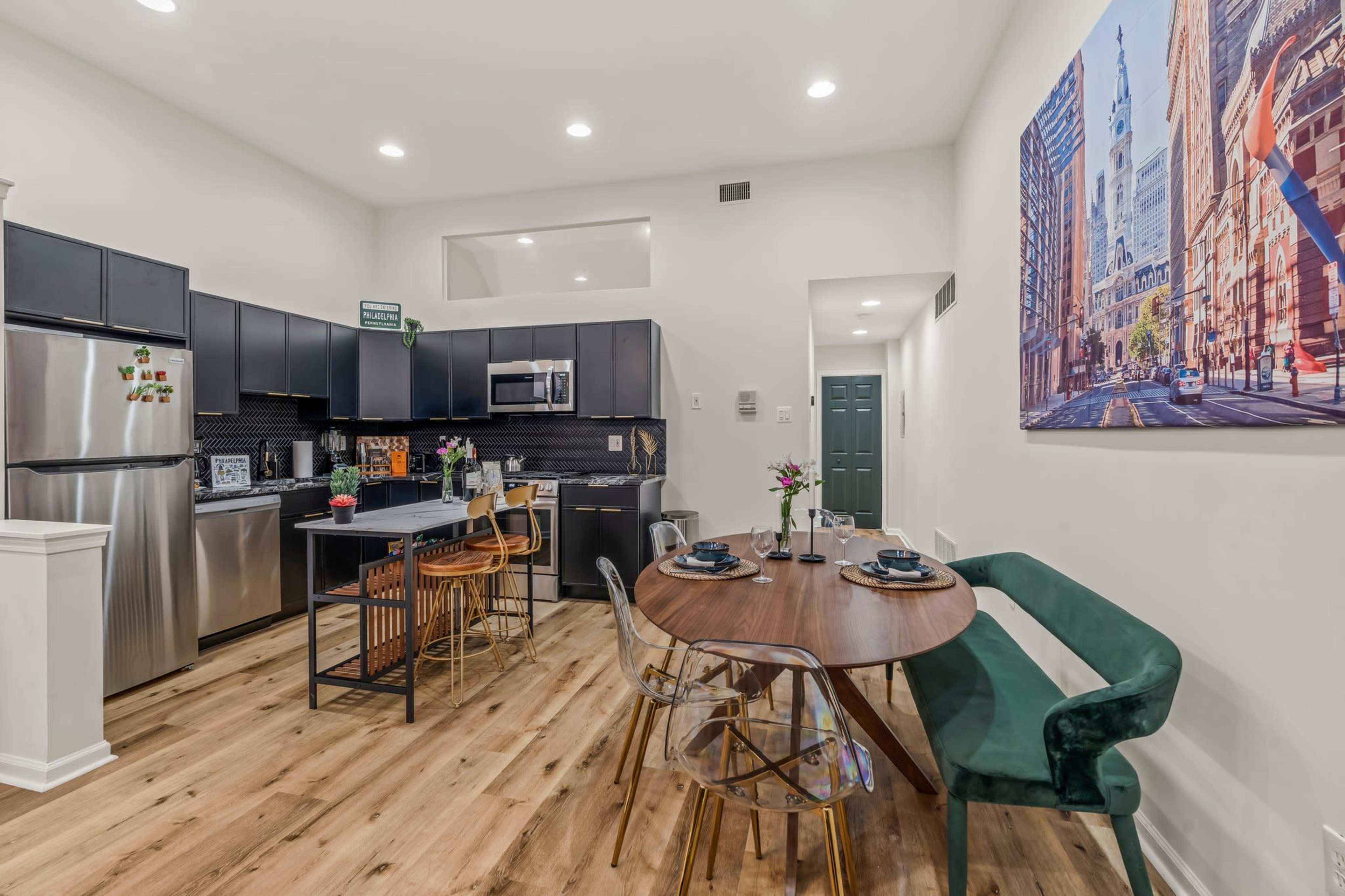 A modern kitchen and dining area features dark cabinetry, stainless steel appliances, a round table set for four, and a large cityscape photograph on the wall.