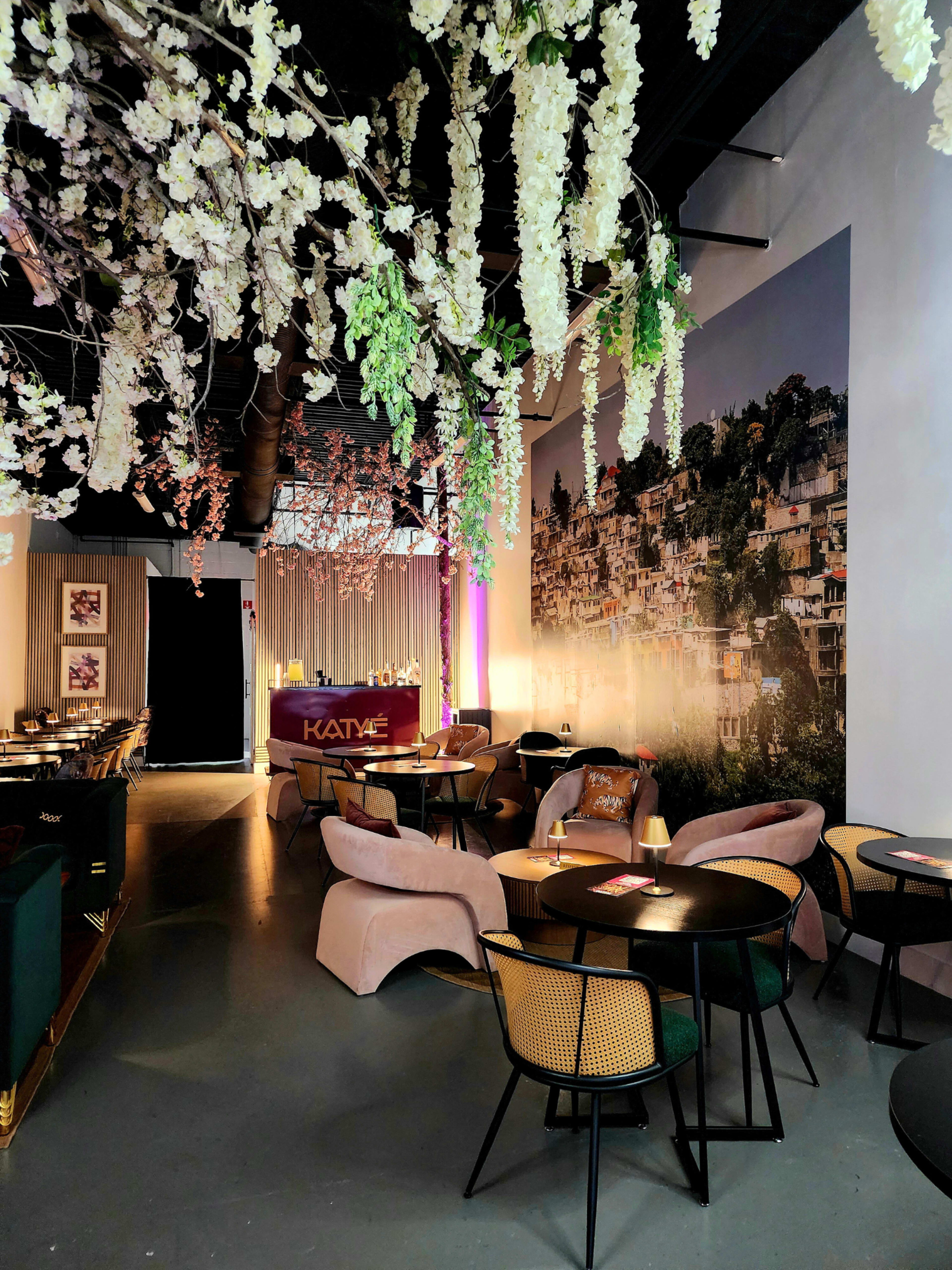 The scene depicts a stylish indoor café with elegant decor, featuring floral arrangements hanging from the ceiling and a mural of a cityscape on the wall.
