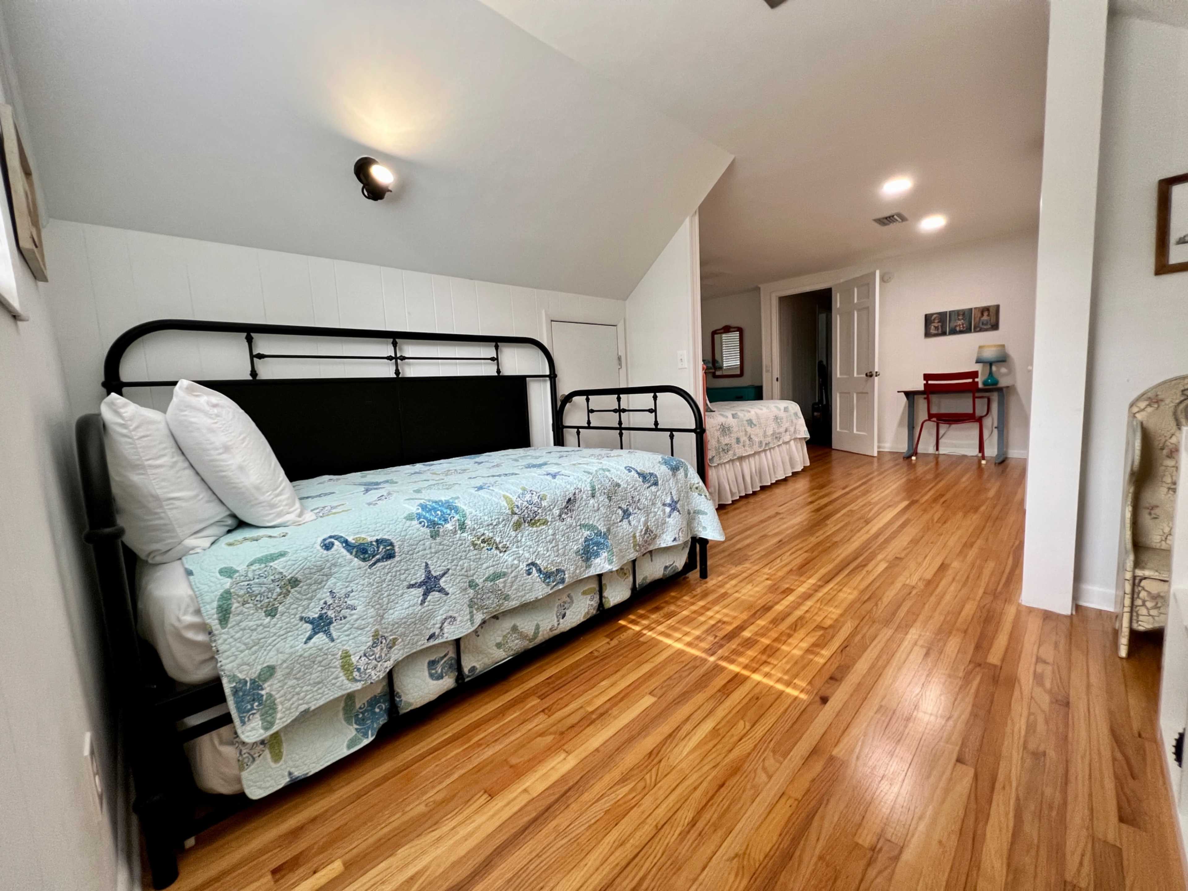 A well-lit room featuring two beds, hardwood flooring, and a small desk and chair against the wall.