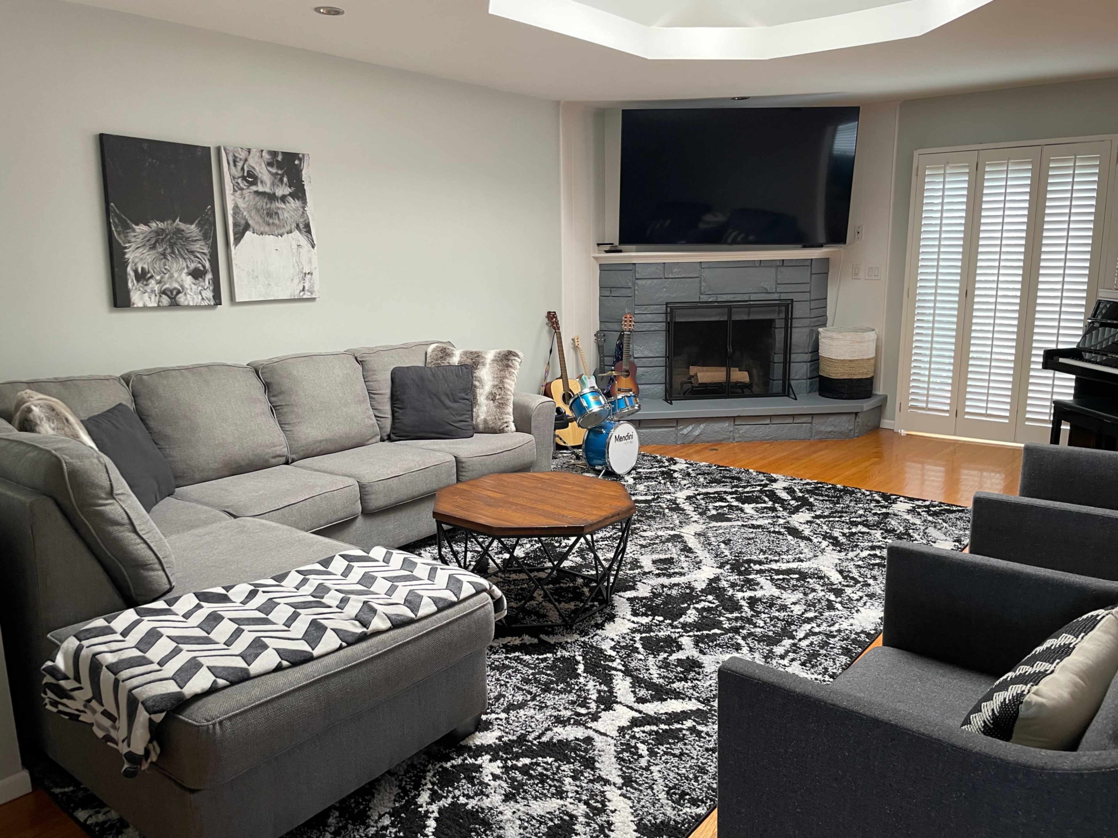 The living room features a gray sectional sofa, a round wooden coffee table, and a fireplace surrounded by a stone facade, with a television mounted above and a patterned area rug on the floor.