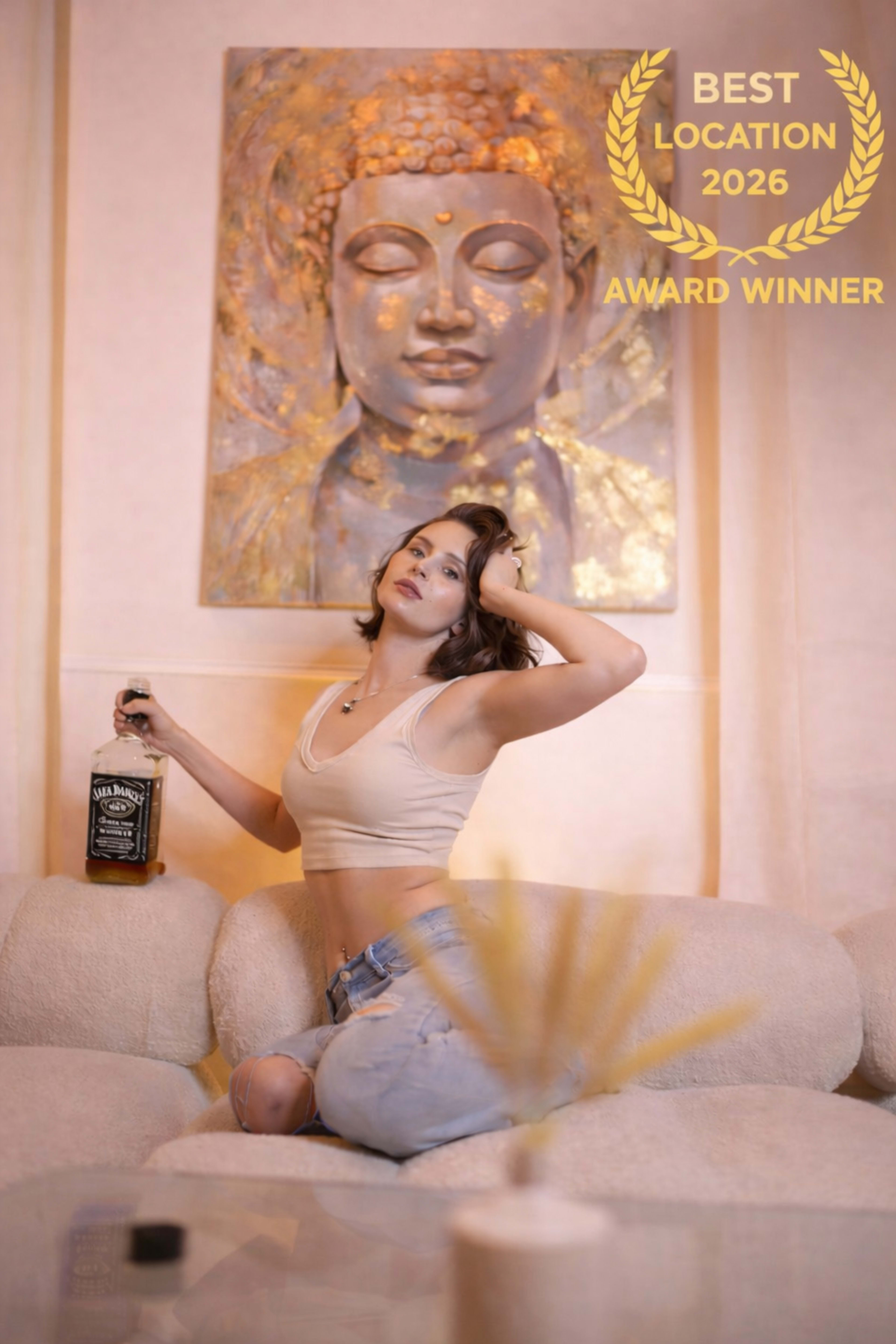 A woman poses on a couch while holding a bottle of whiskey in front of a large Buddha mural.