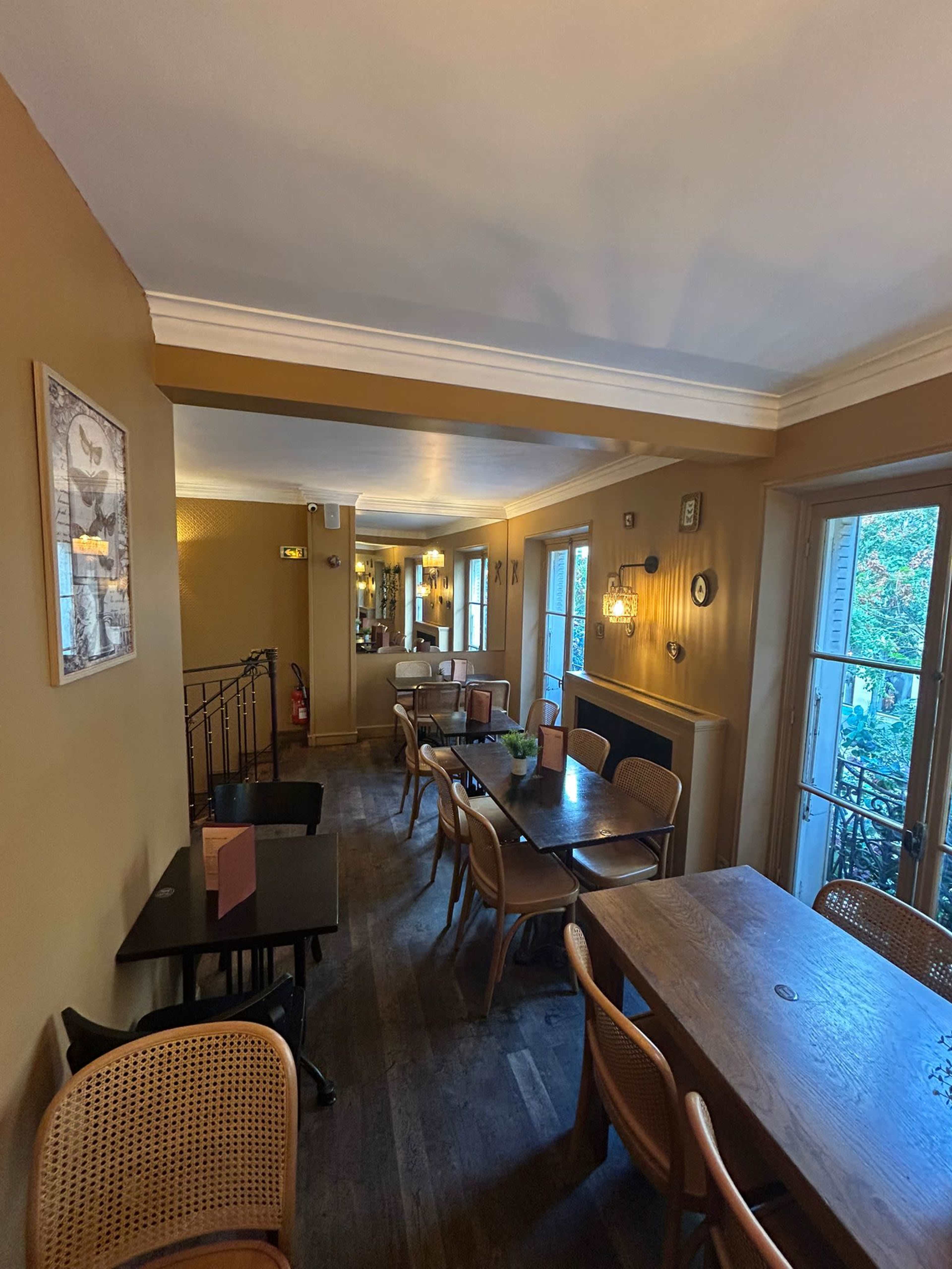 The image shows a warmly lit dining area in a restaurant with wooden tables and chairs, along with decorative wall art and windows offering a view outside.