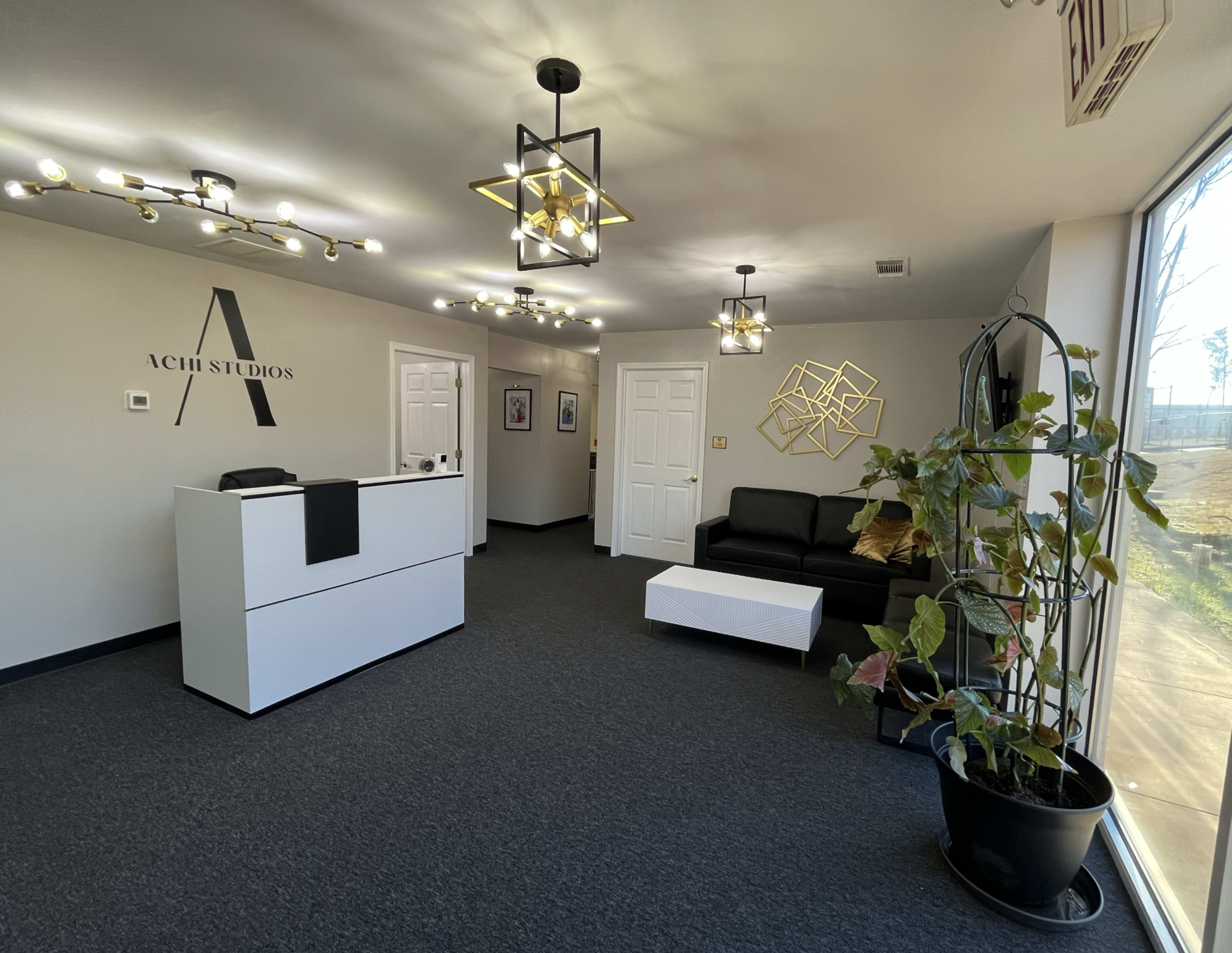 The image shows a modern reception area featuring a sleek white desk, a black sofa, a decorative plant, and contemporary lighting.