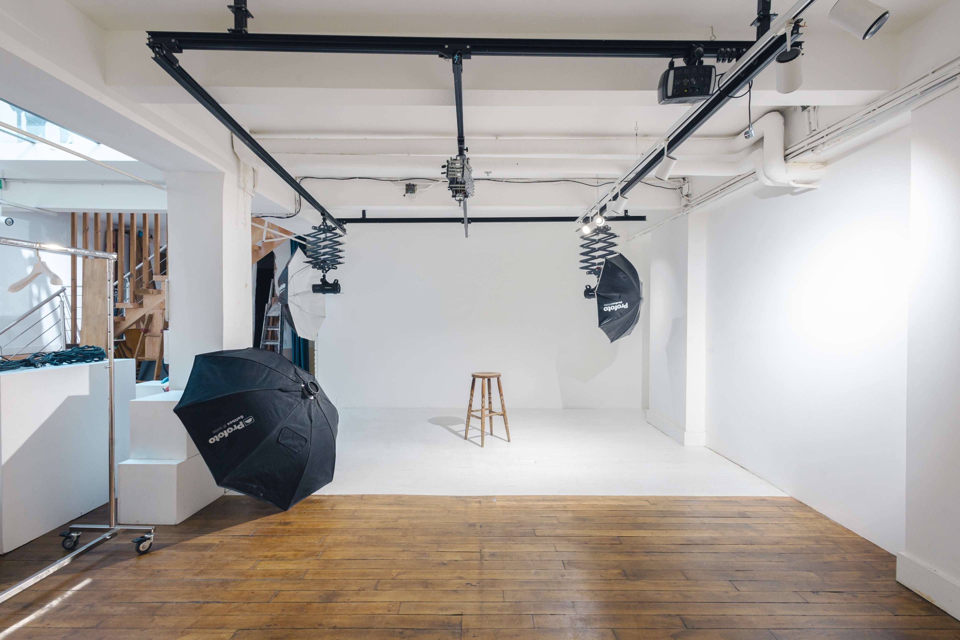 A photography studio features a white backdrop, a wooden stool, and several light umbrellas positioned around the space.