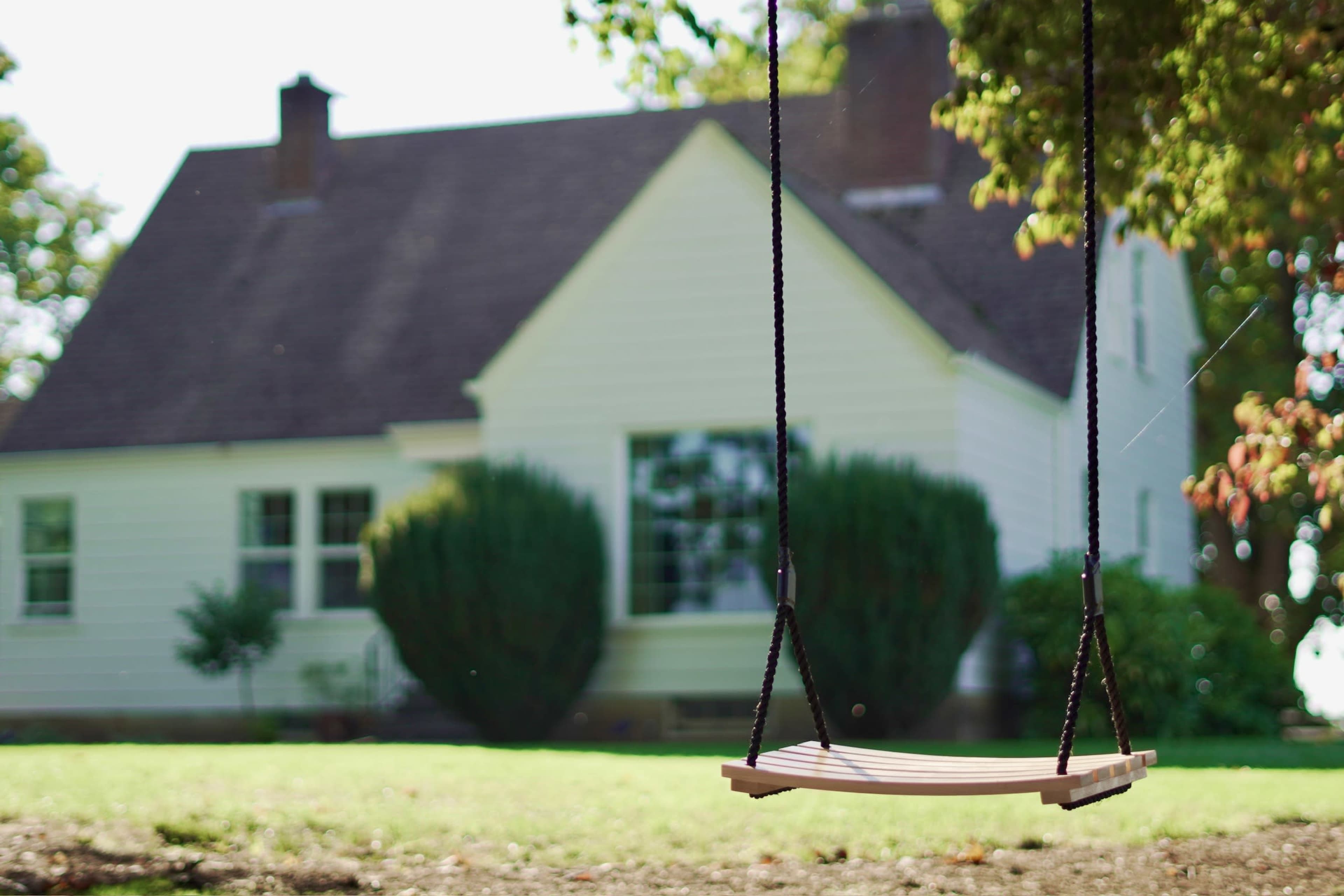 A wooden swing hangs from a rope in the foreground, with a house and garden visible in the background.