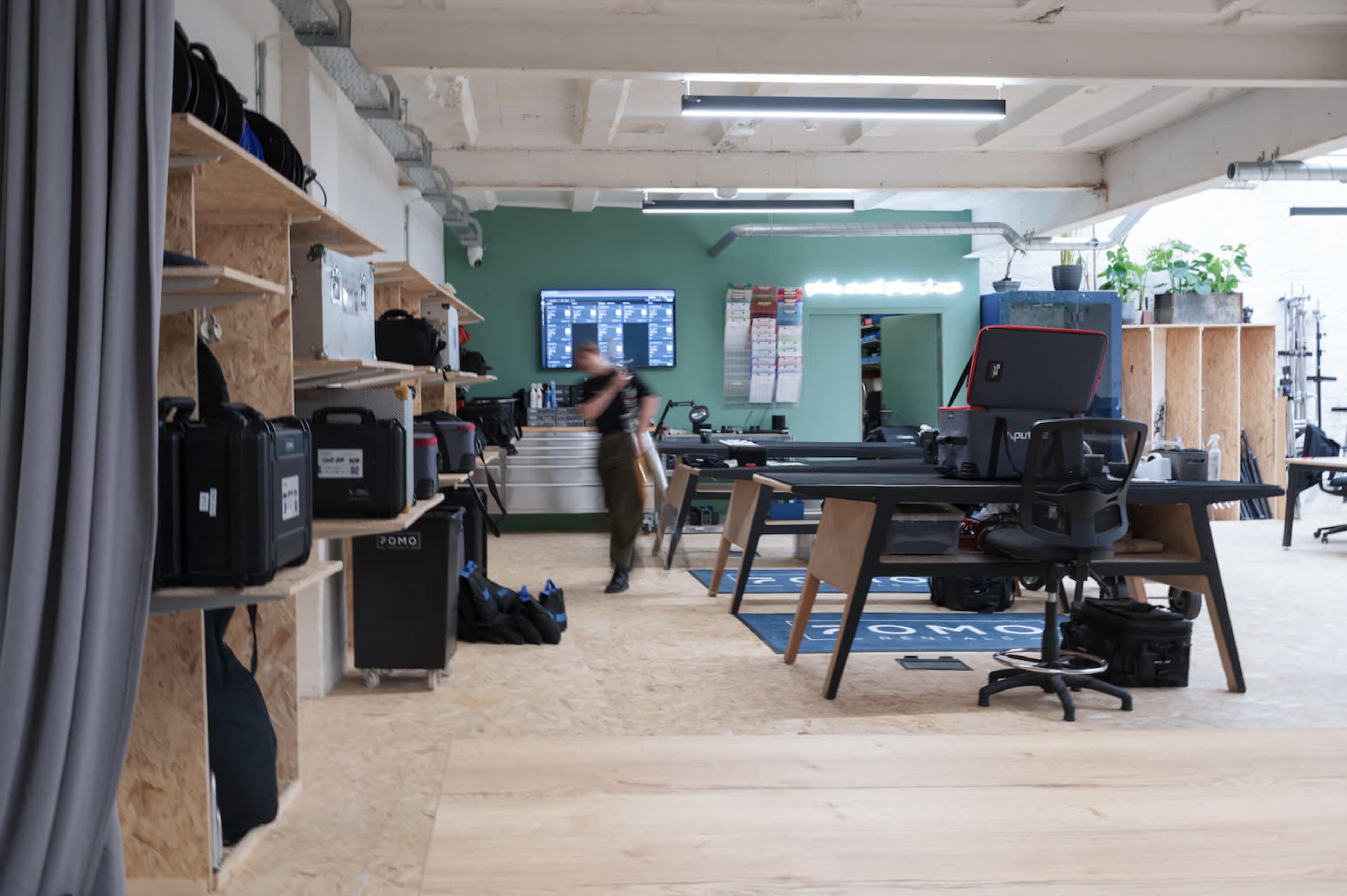 A spacious workspace with wooden shelving holding various gear, a large screen on the wall, and a desk area with a chair in a modern, industrial-style setting.