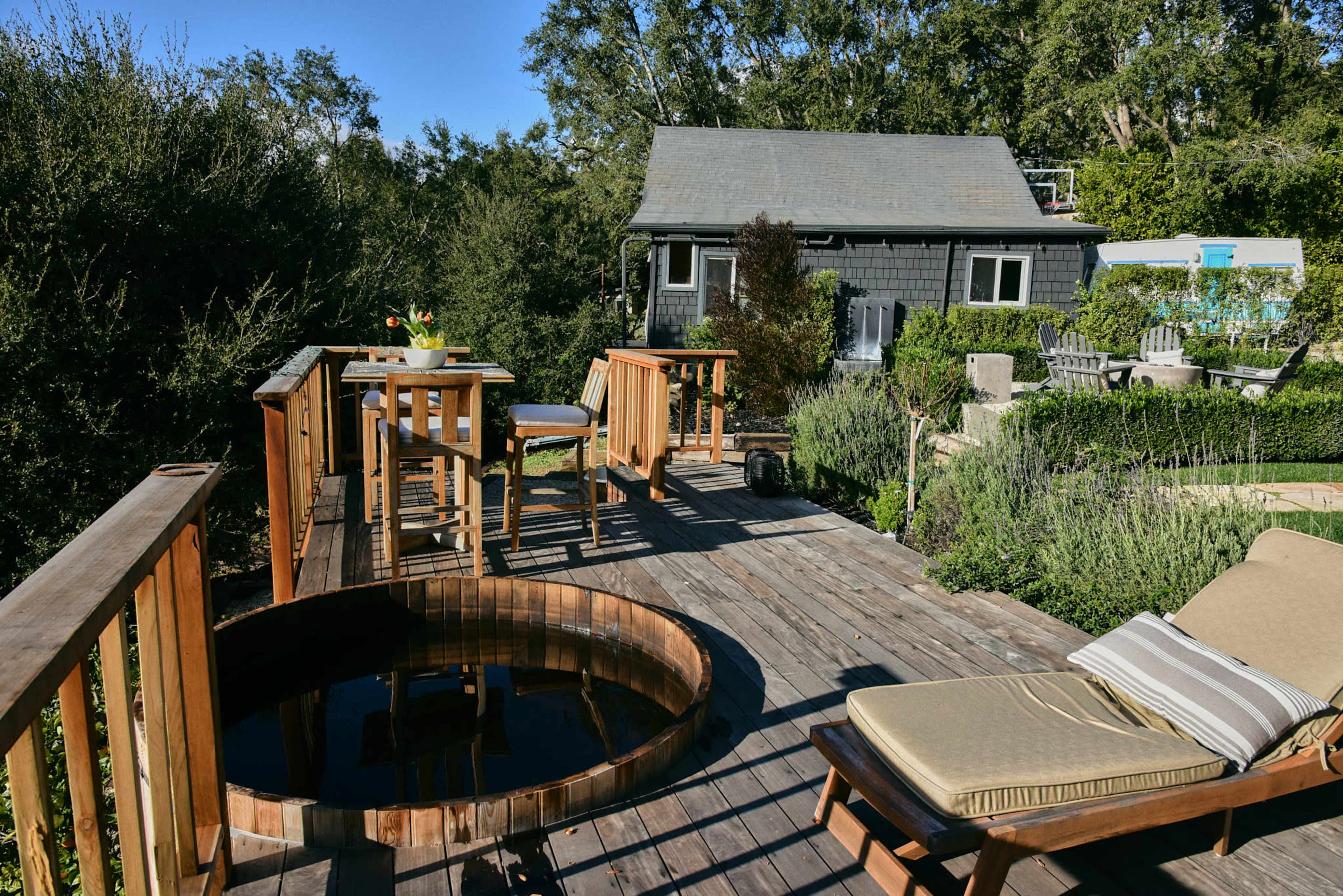 A wooden deck features a hot tub, a set of outdoor furniture, and a small house in the background surrounded by greenery.