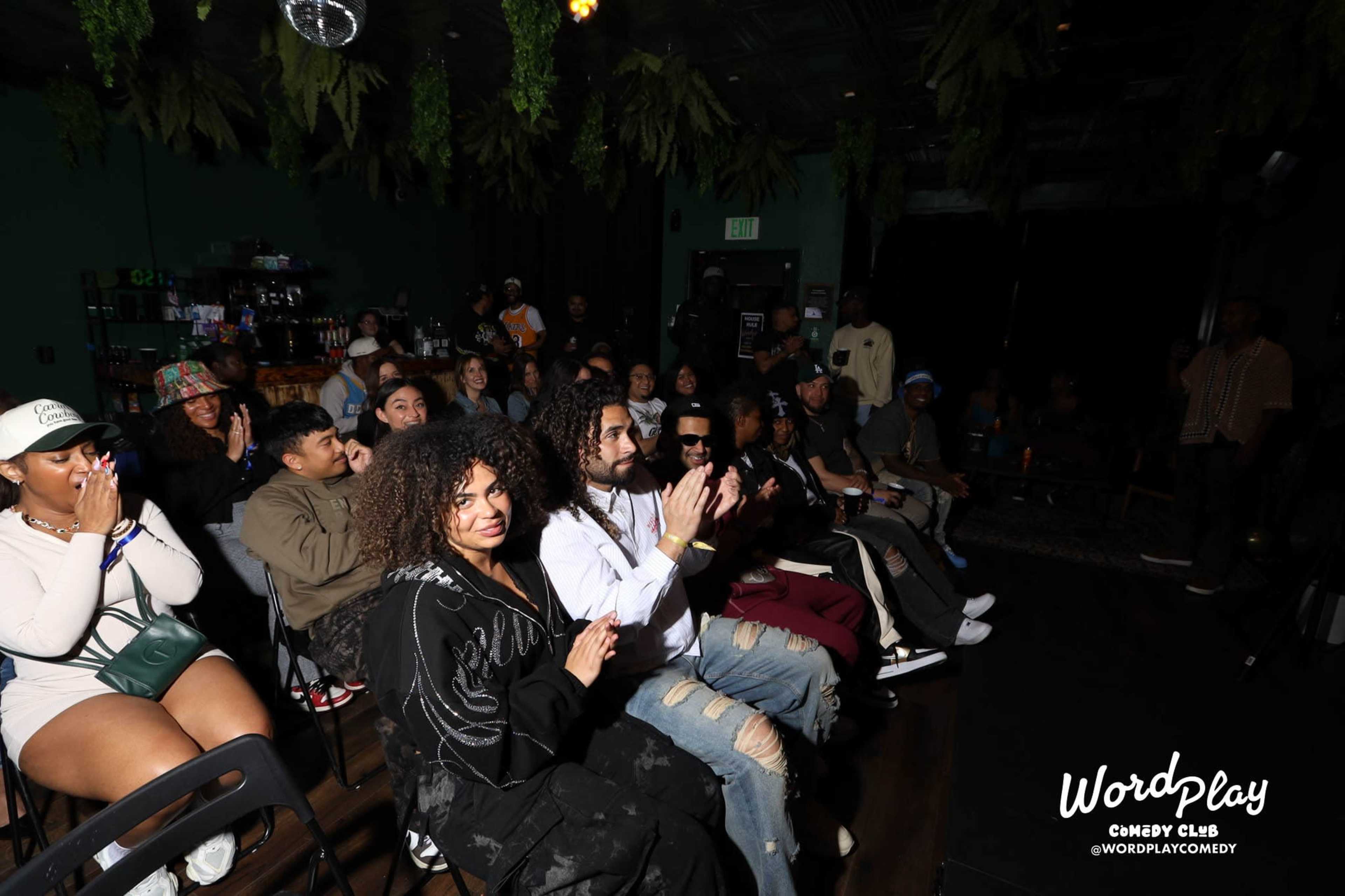 A crowd of people sits in a dimly lit comedy club, some smiling and clapping, while others observe the stage.