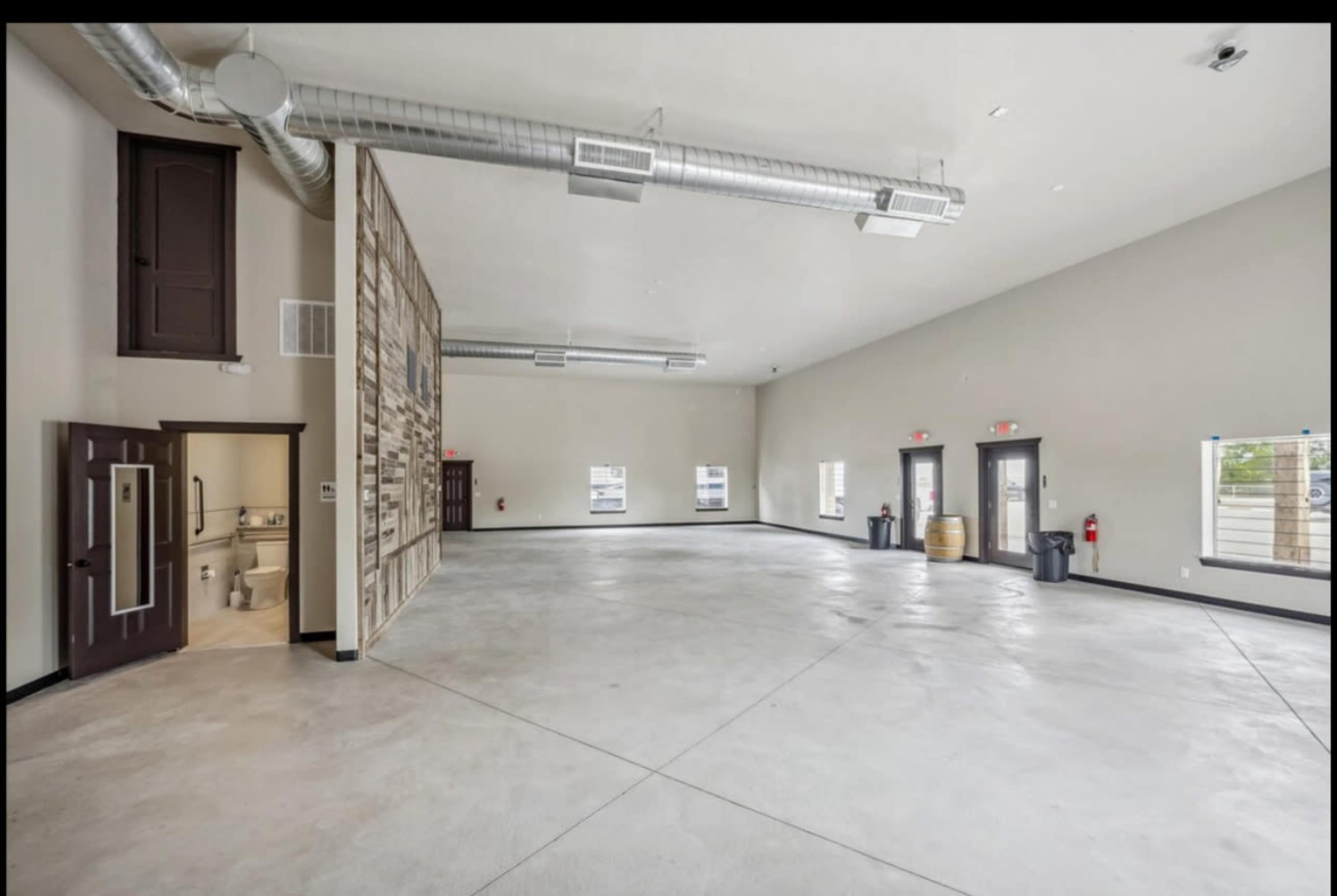 The image shows a spacious, empty room with polished concrete floors, high ceilings, and large windows, featuring a small restroom area visible to the left.
