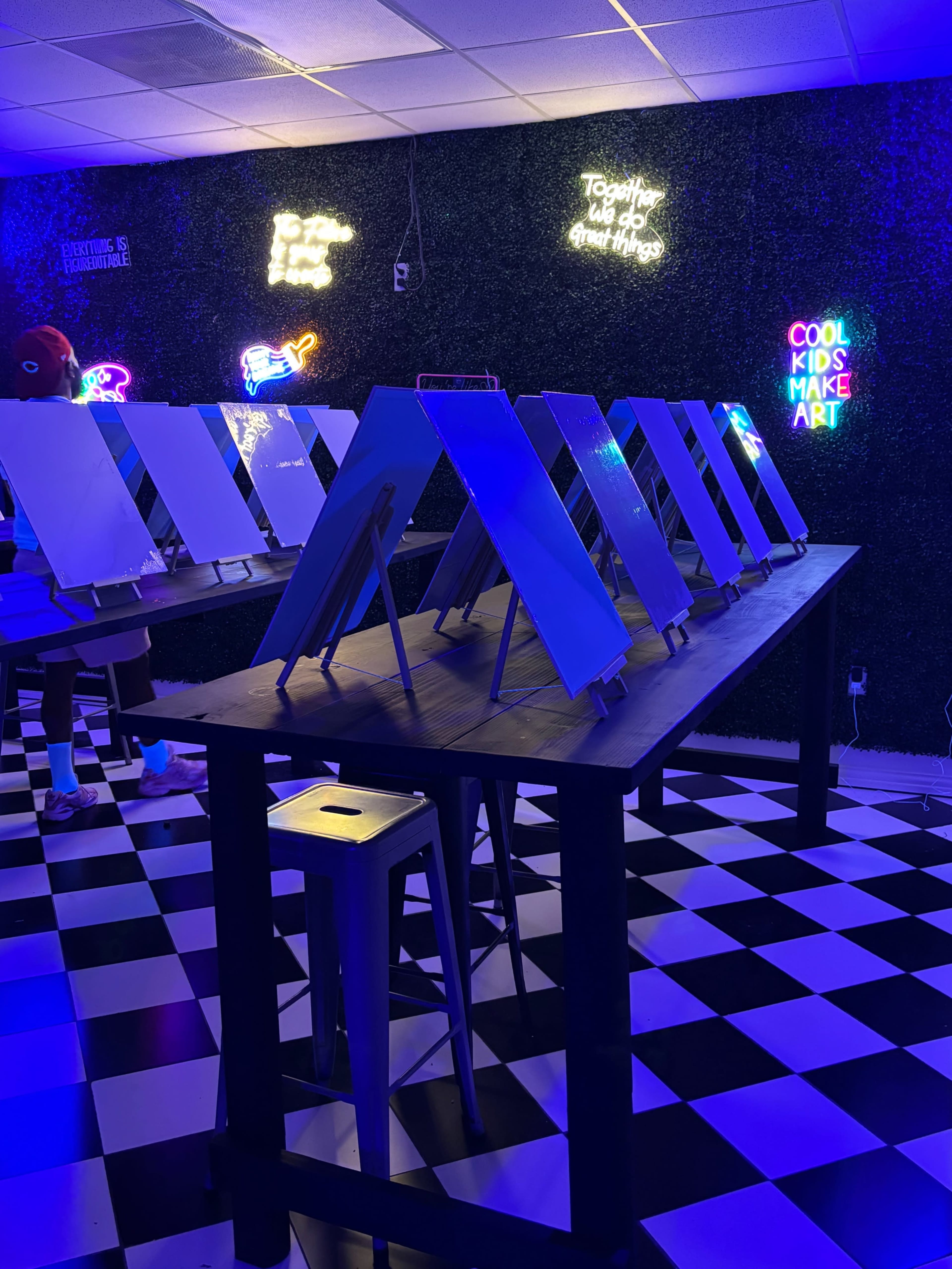 The image shows a brightly lit art studio with several easels lined up on a black and white checkered floor, accompanied by colorful neon signs on the wall.