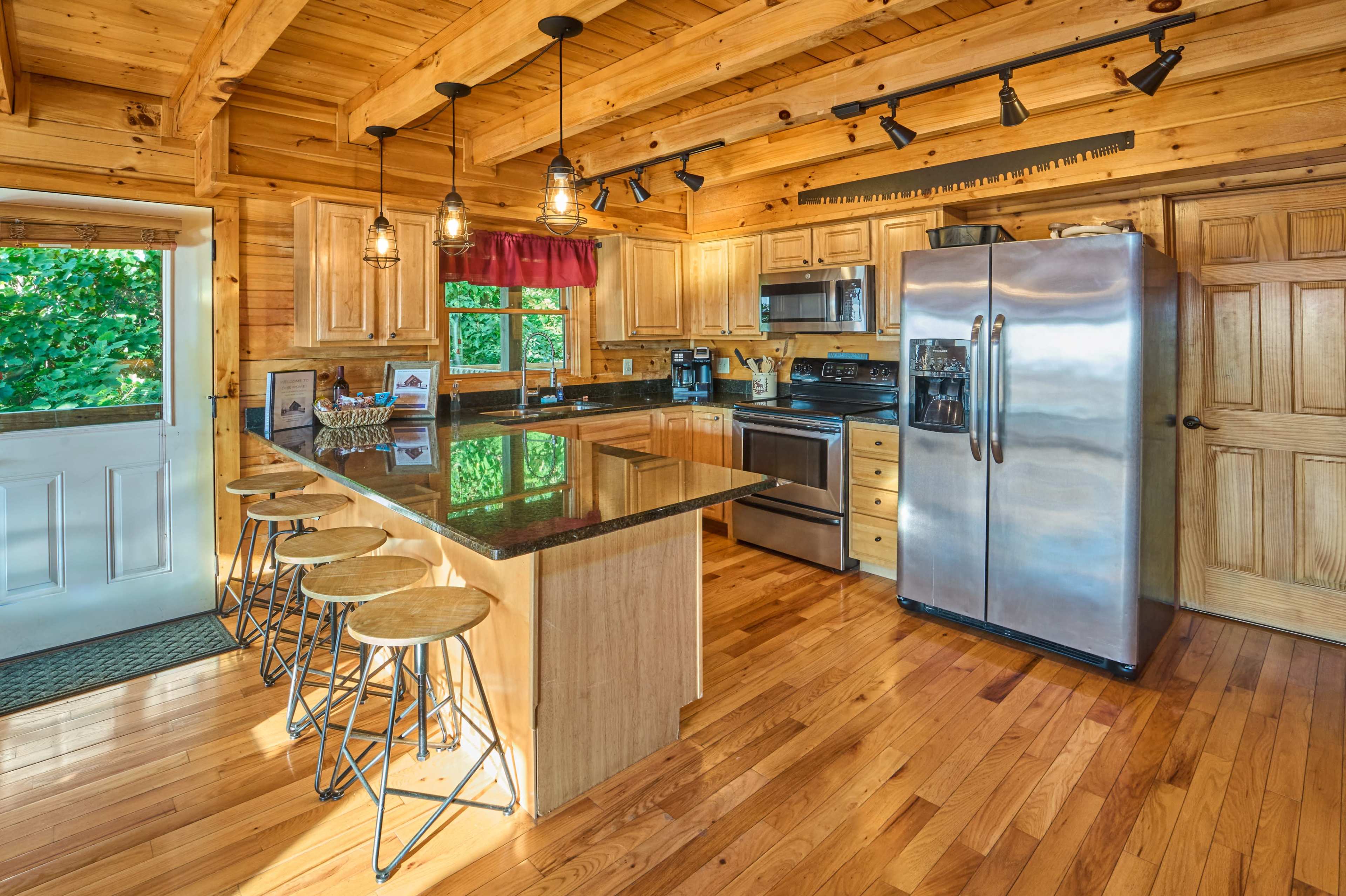 The image depicts a wooden kitchen with a large island featuring three bar stools, stainless steel appliances, and natural light coming from a door and window.