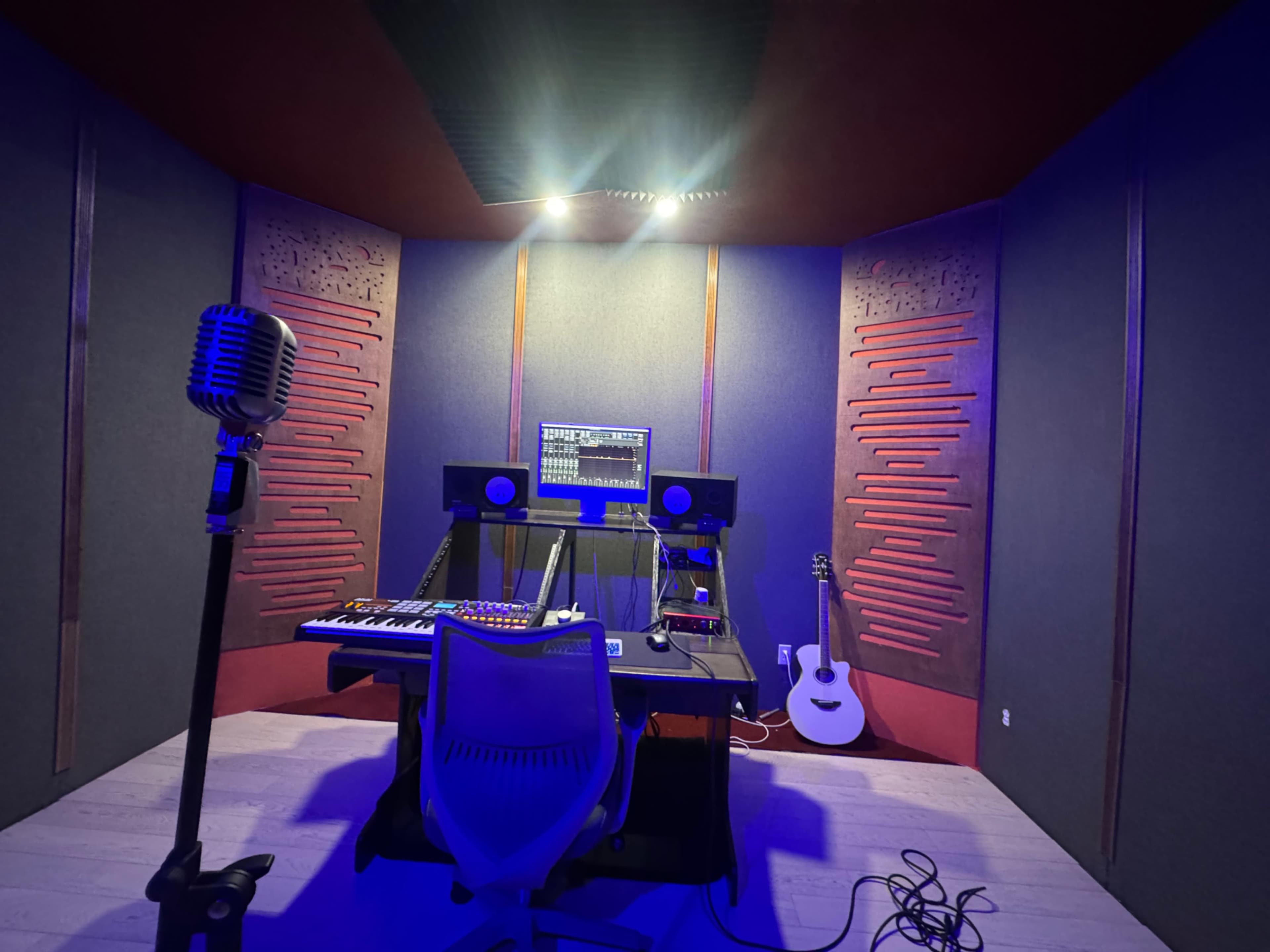 The image shows a music studio with soundproofed walls, a microphone in the foreground, a keyboard and mixer on the desk, and an acoustic guitar leaning against the wall.