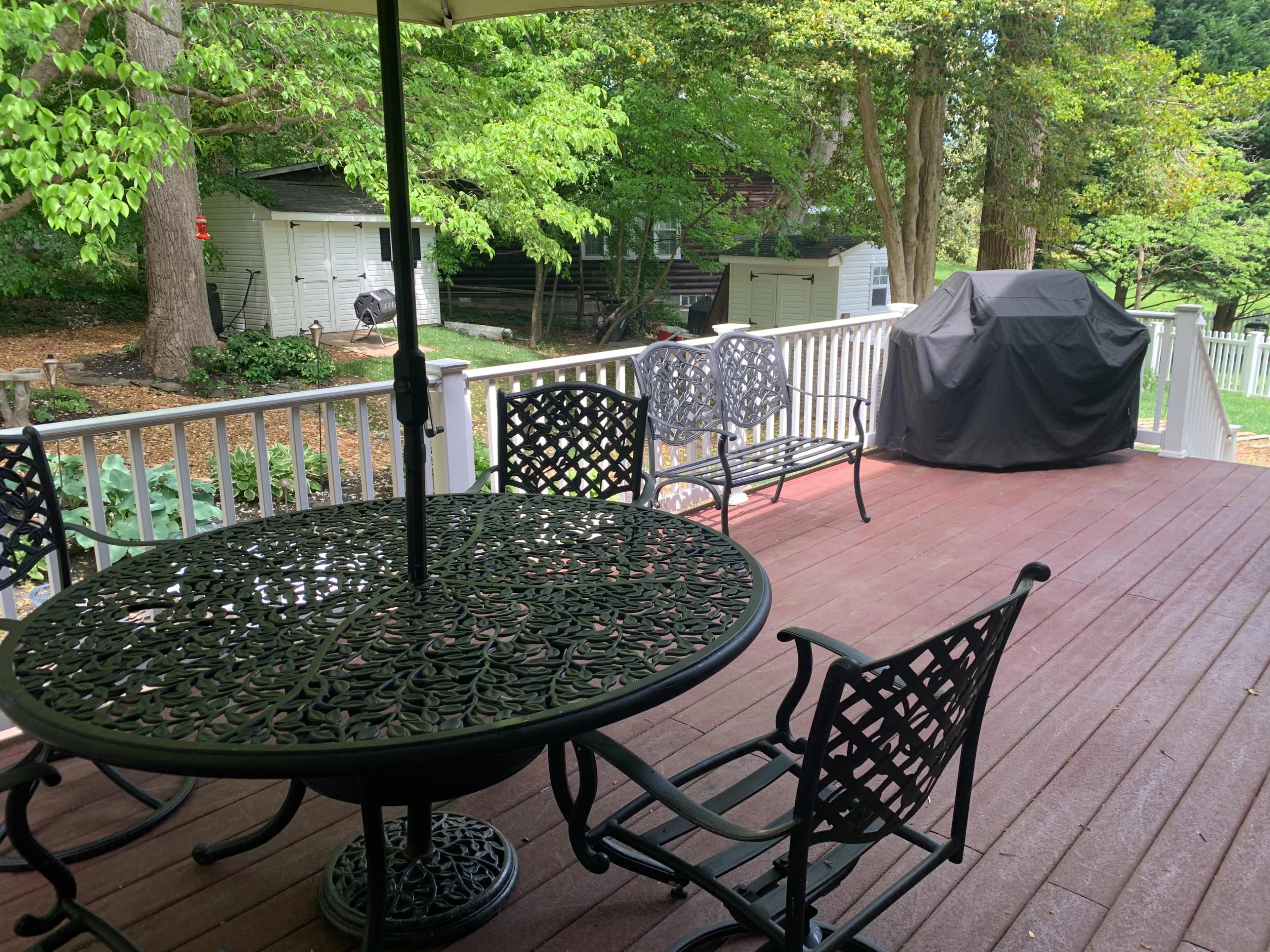 A patio area features a round metal table with chairs, an umbrella, and a grill, surrounded by lush greenery.