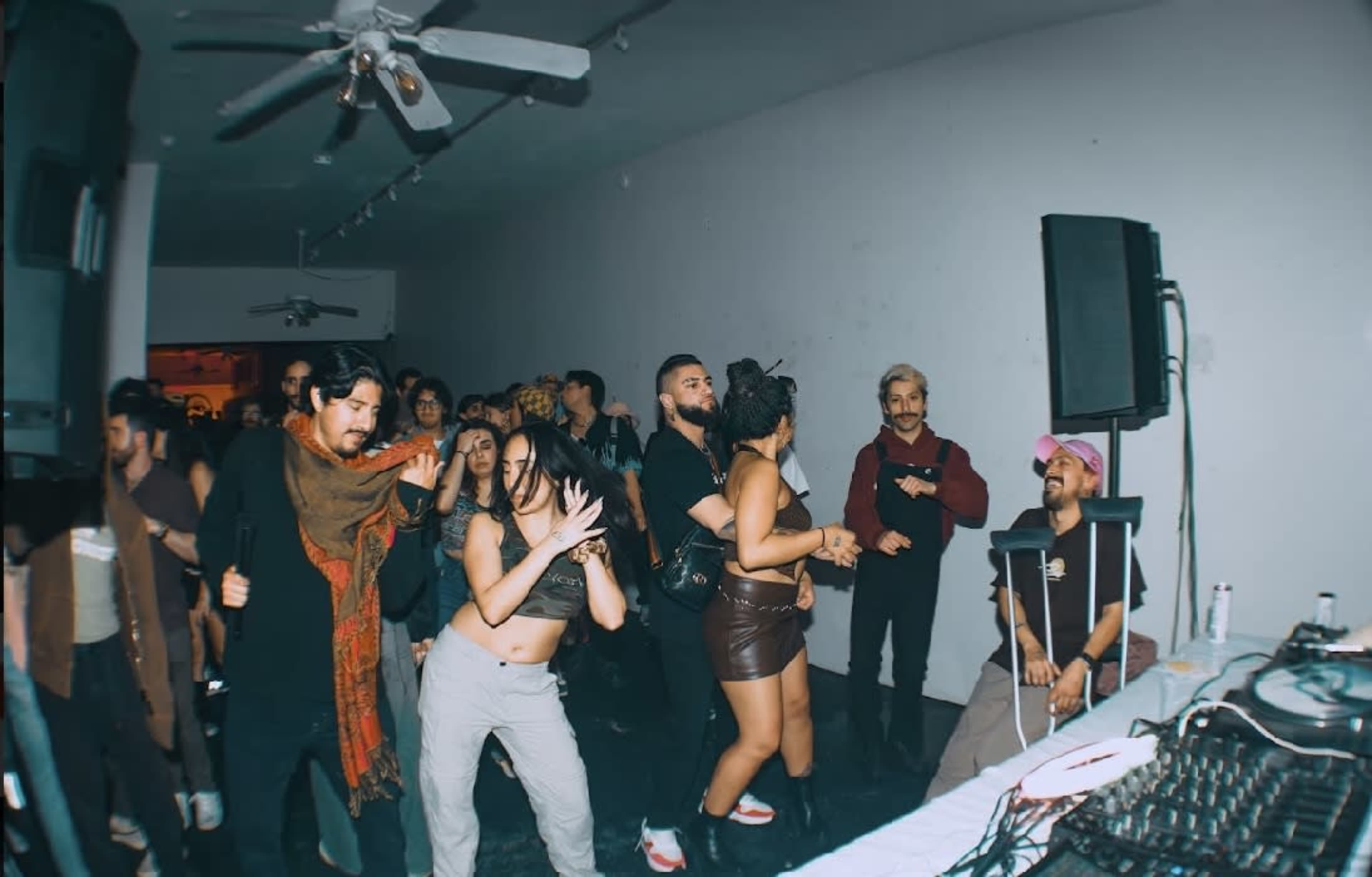 A group of people dances in a dimly lit space with a DJ setup and sound equipment in the foreground.