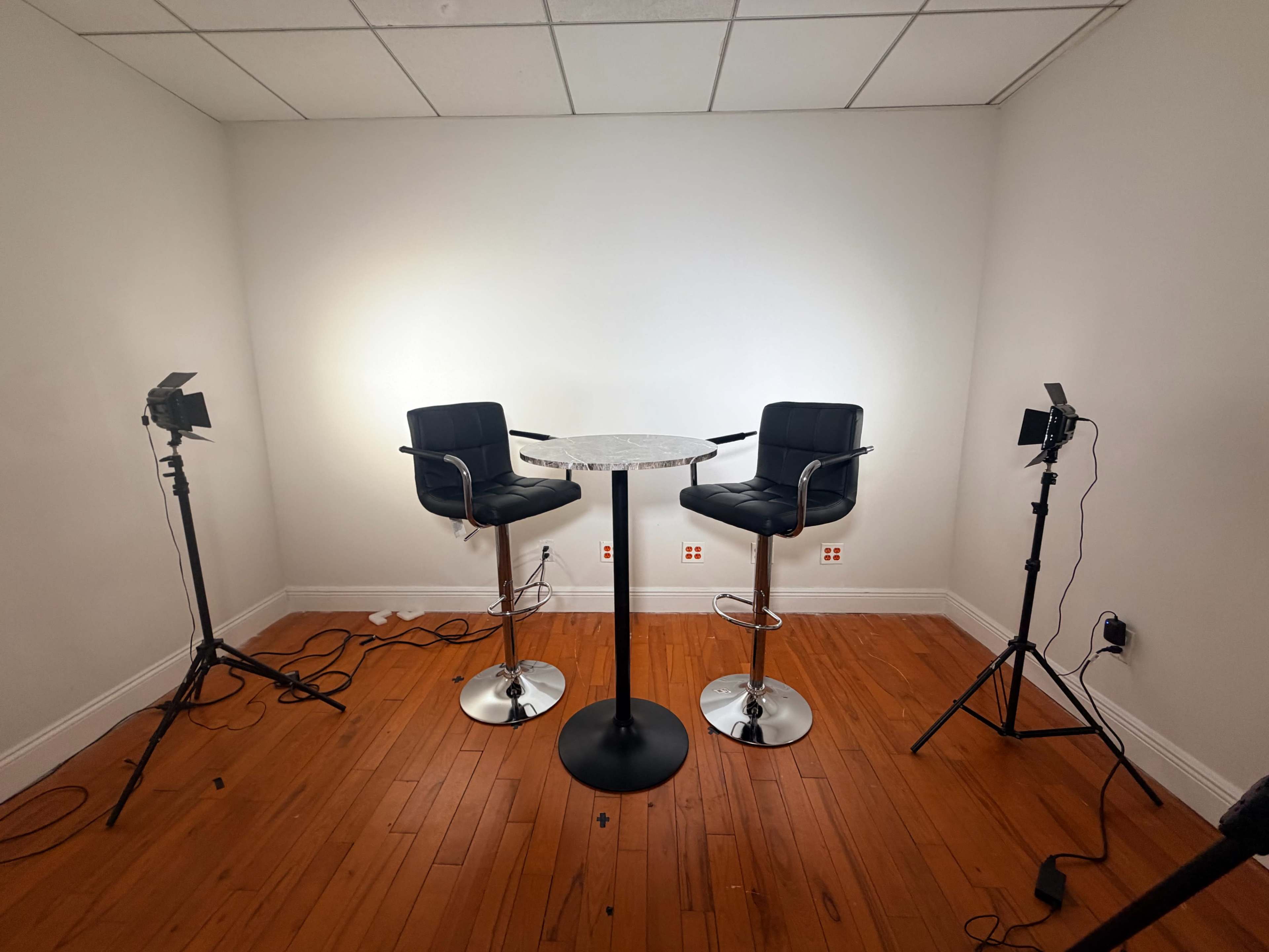 A small room features a circular table and two black bar stools, illuminated by two light stands on either side.