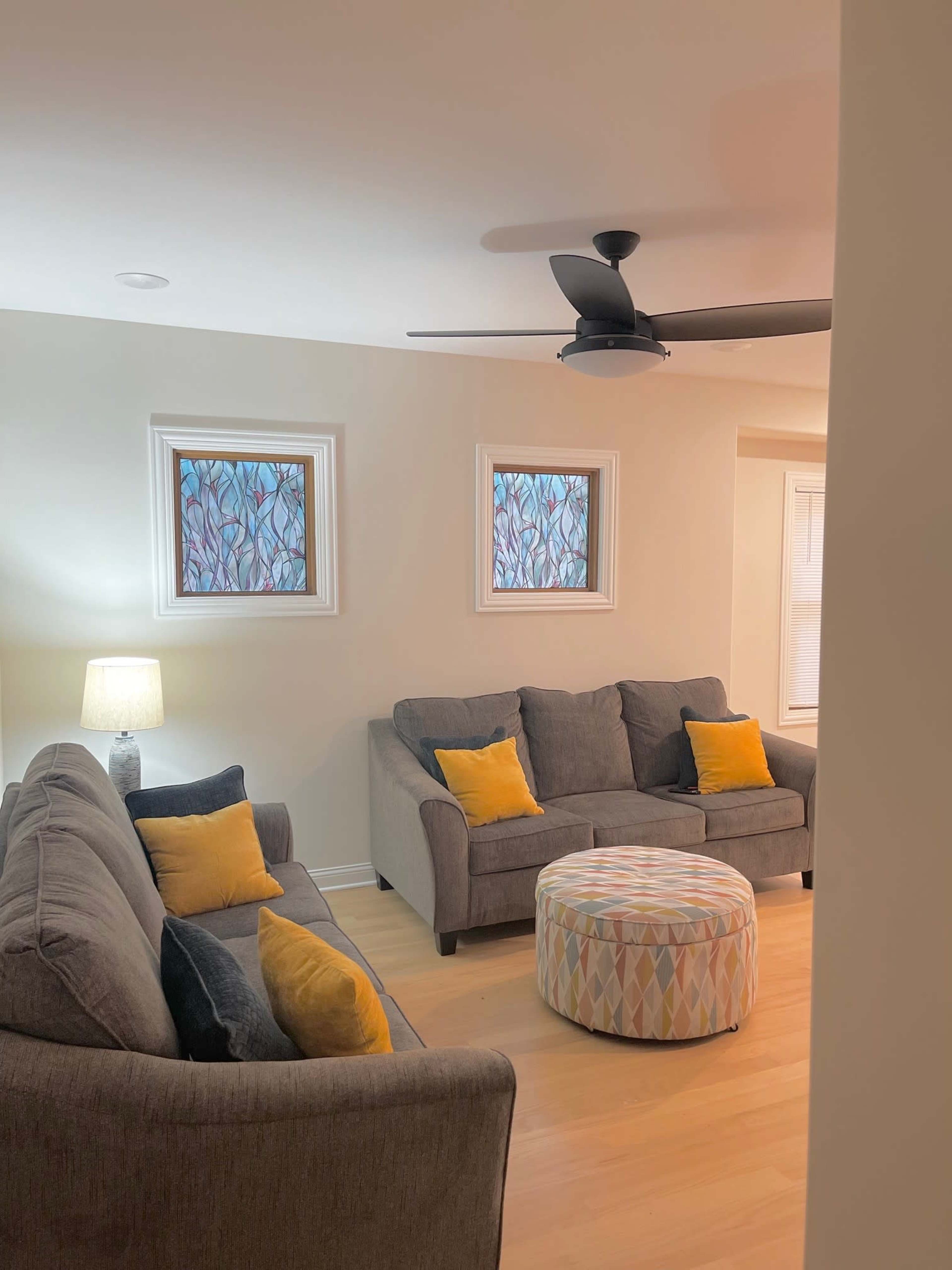 The living room features two gray sofas with yellow and black cushions, a round ottoman, and colorful stained glass windows.