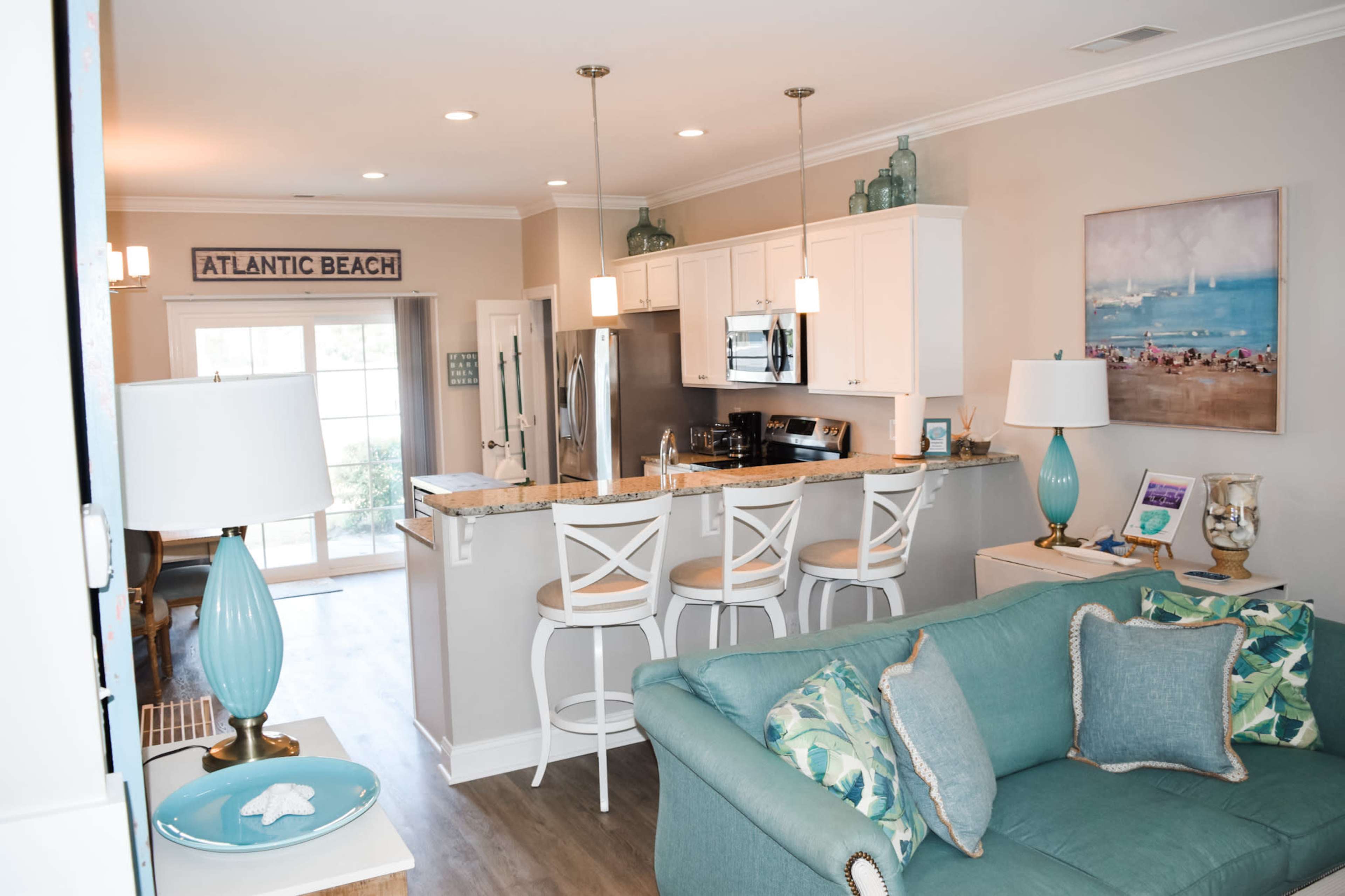 A modern, open-concept kitchen and living area with white cabinets, a granite countertop, and bar stools, accented by decorative lighting and coastal-themed decor.
