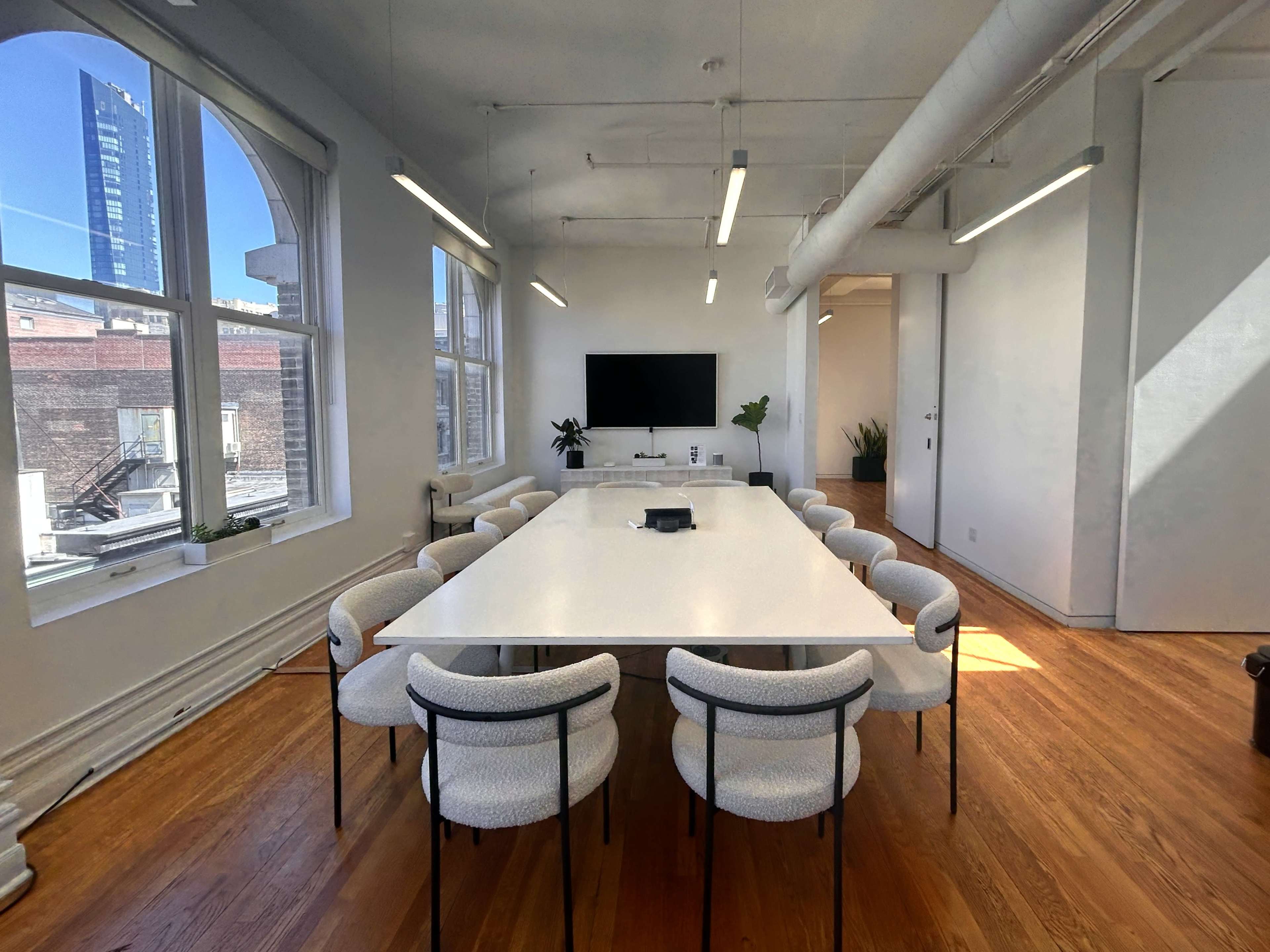 A modern conference room with a large white table surrounded by twelve chairs, windows on one side, and a television mounted on the wall.