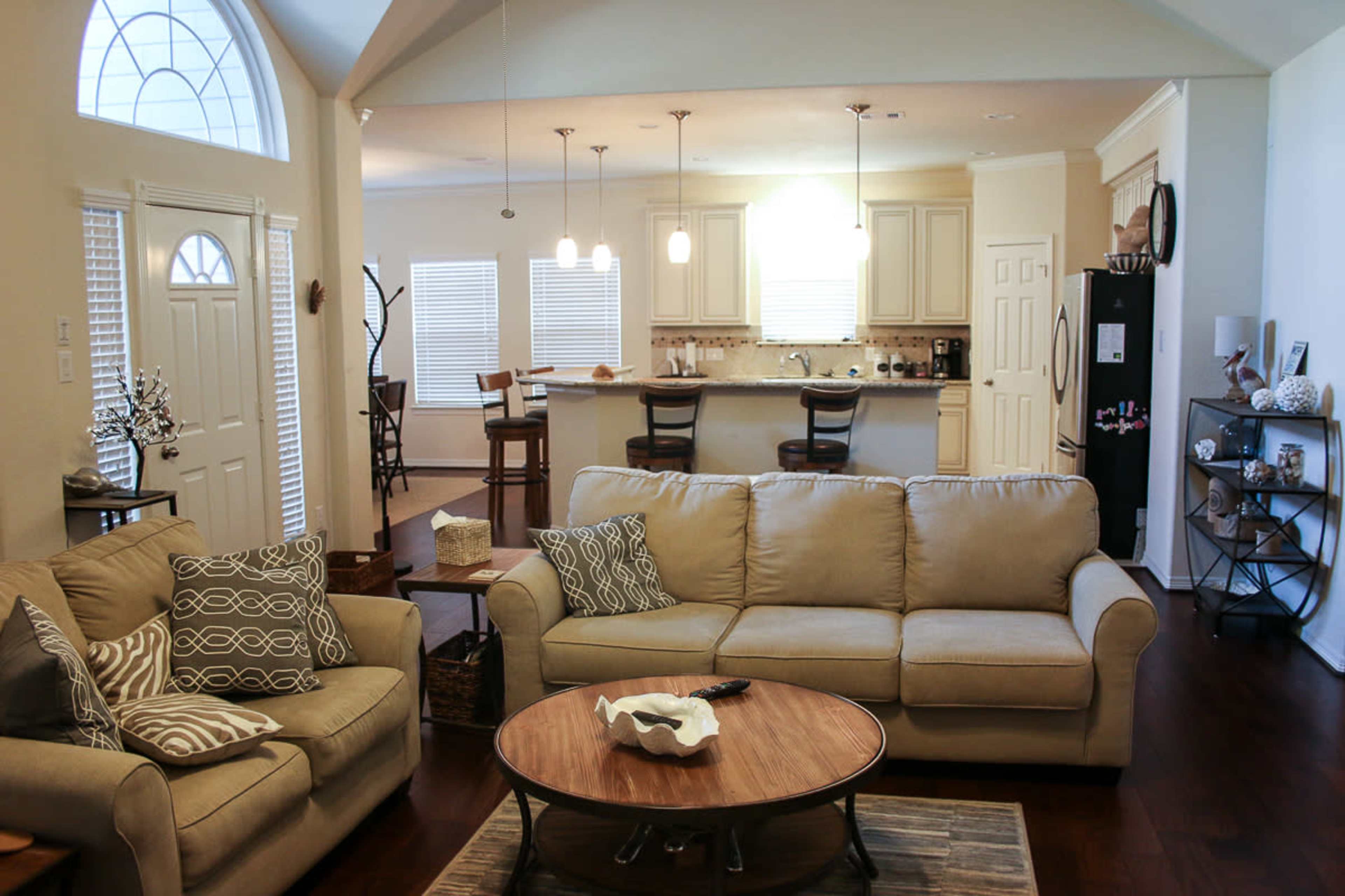 The image shows a spacious living room with a beige sofa, a coffee table, and an open layout leading to a kitchen area with white cabinetry and light fixtures.