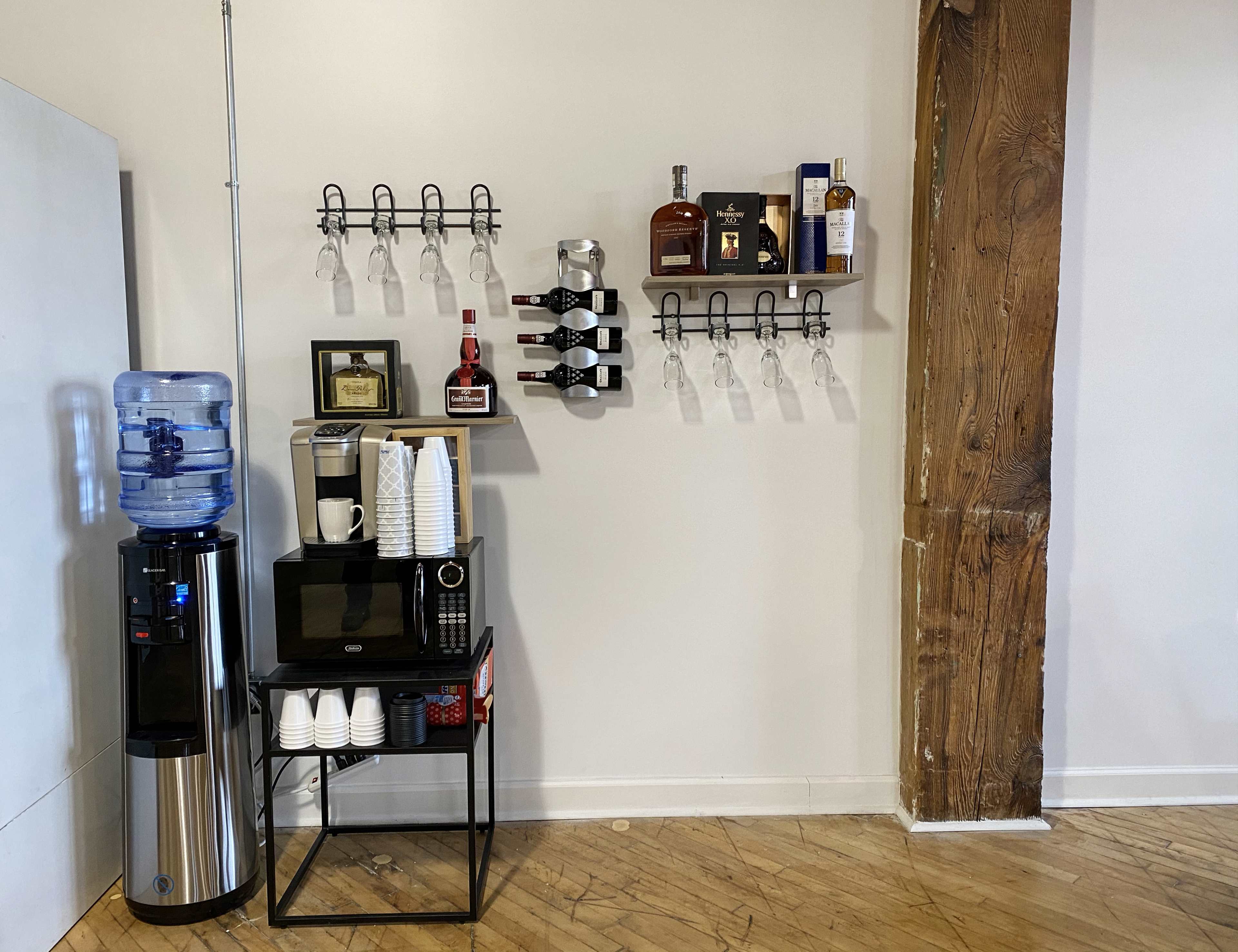 The image shows a beverage station featuring a water cooler, a microwave, stacked cups, and shelves displaying various bottles and wine glasses against a wooden pillar.