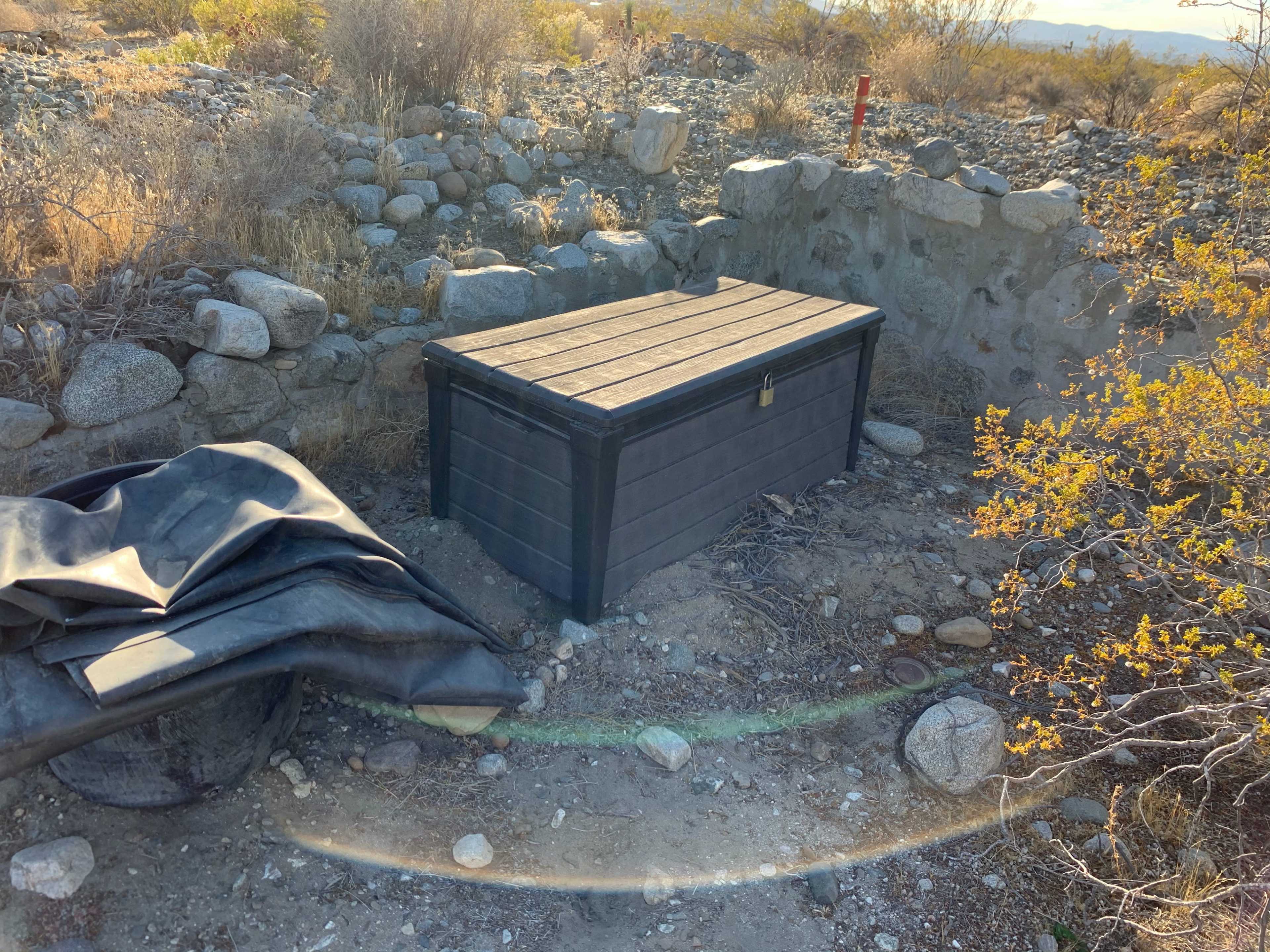 A wooden storage box is placed on rocky ground surrounded by sparse vegetation and stone structures.