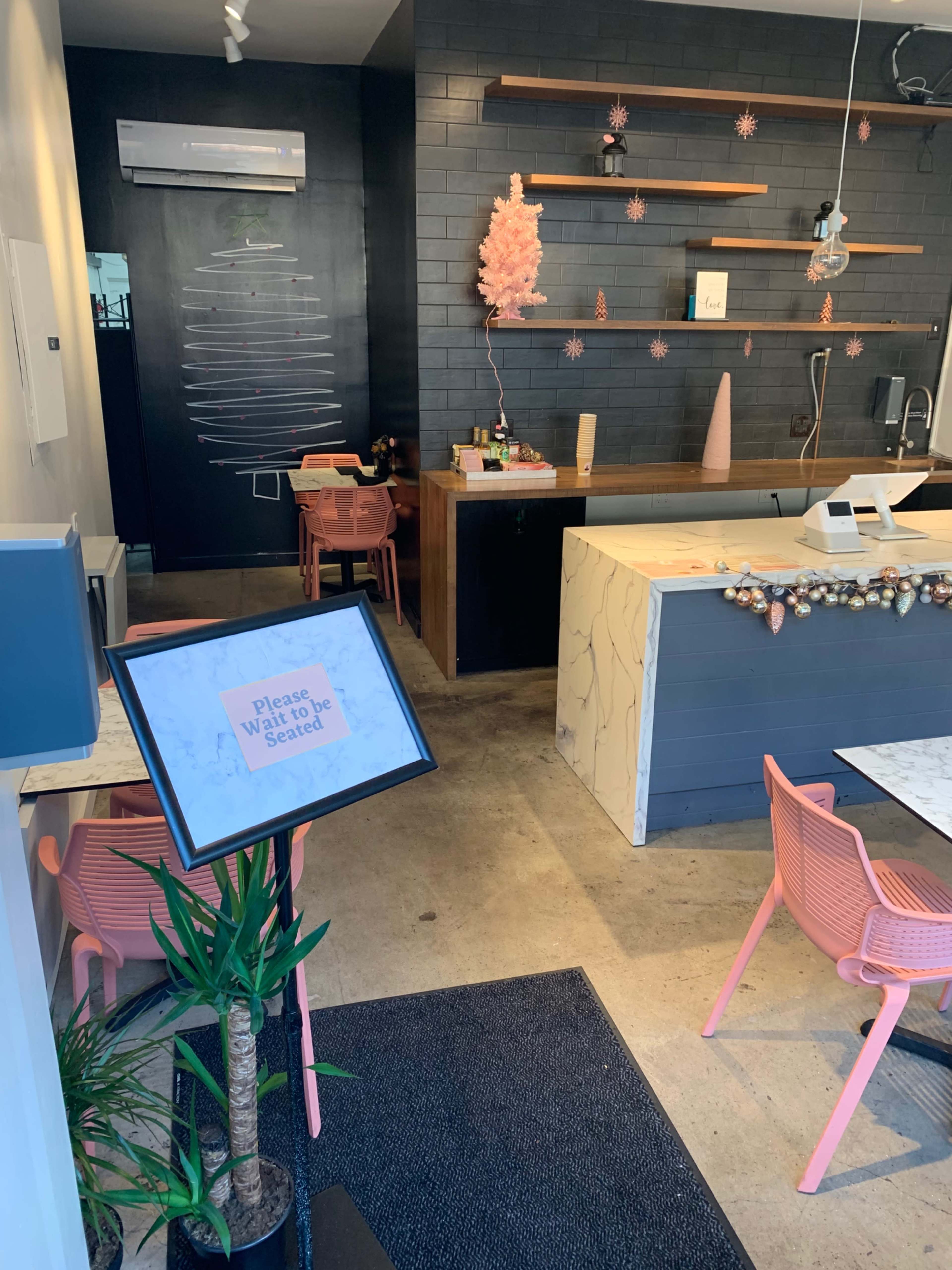 A modern café interior featuring a reception area with a sign requesting patrons to wait to be seated, along with pink chairs, a marble counter, and minimalist decor.