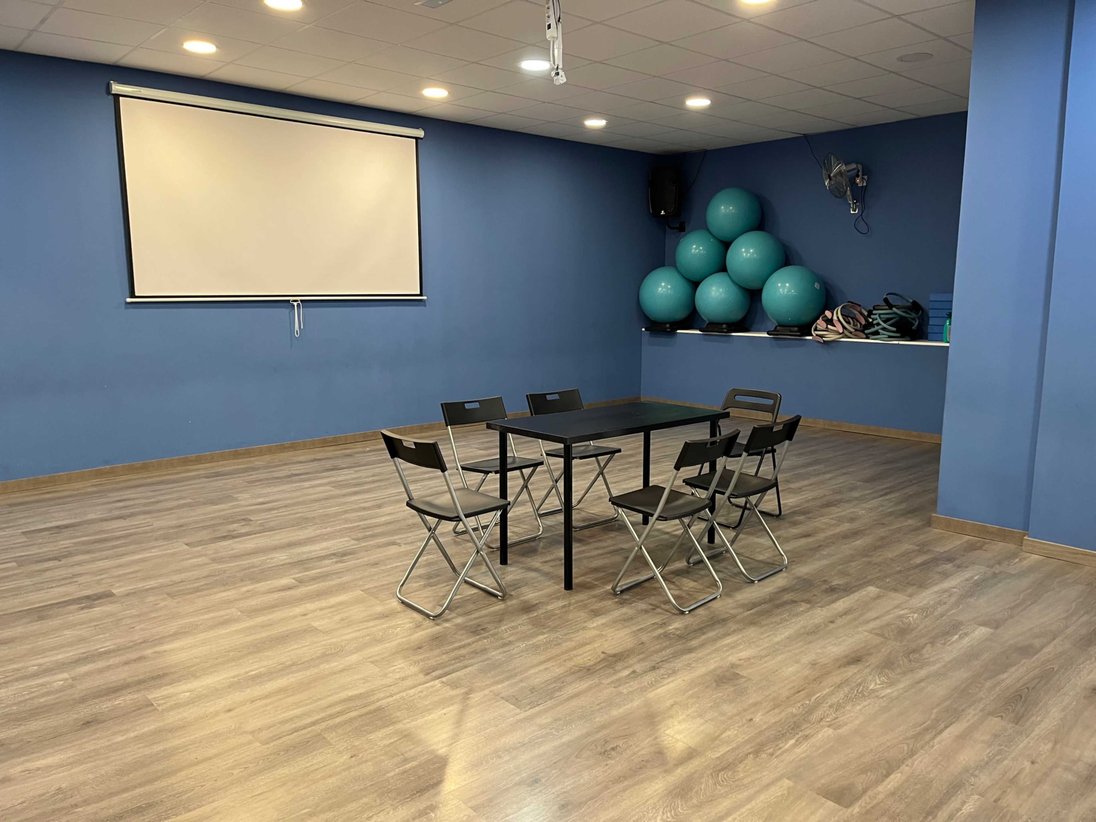 A meeting room features a rectangular table surrounded by six folding chairs, with a projector screen mounted on a blue wall and exercise balls stored nearby.